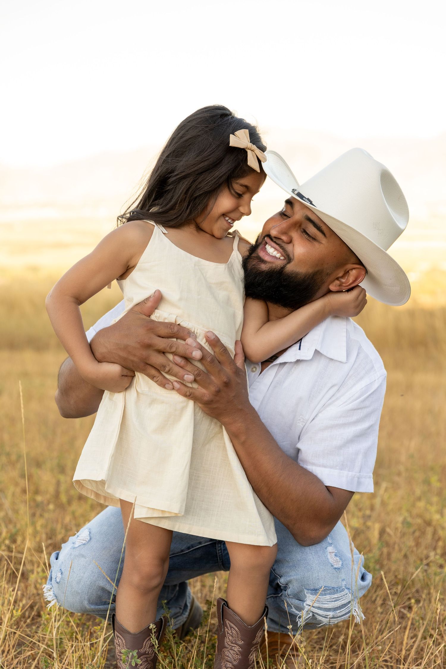 A bearded man in a cowboy hat kneels in a golden field, joyfully embracing his young daughter in a cream dress.