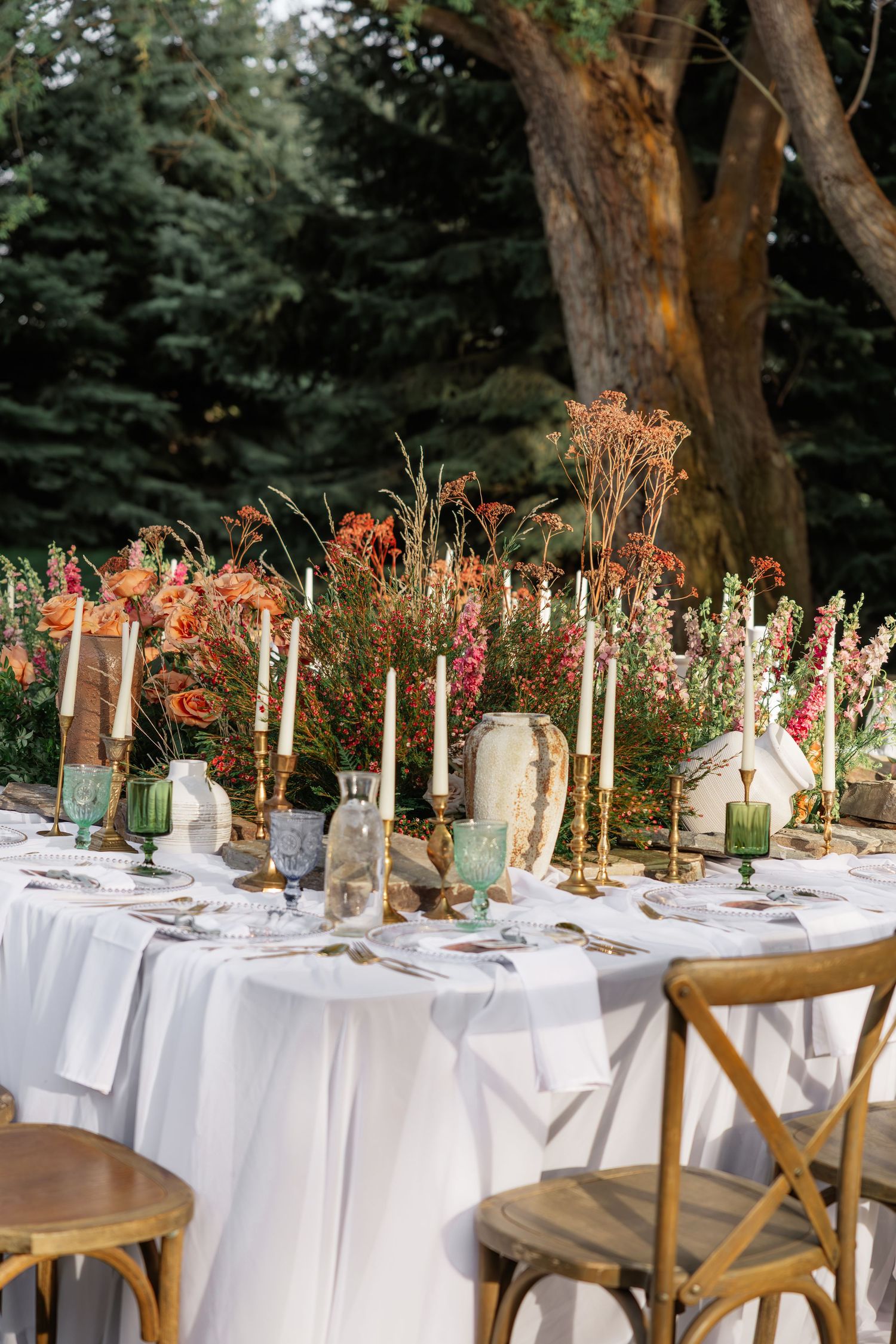 Elegant outdoor wedding reception table with candles, floral centerpieces, and green glassware.