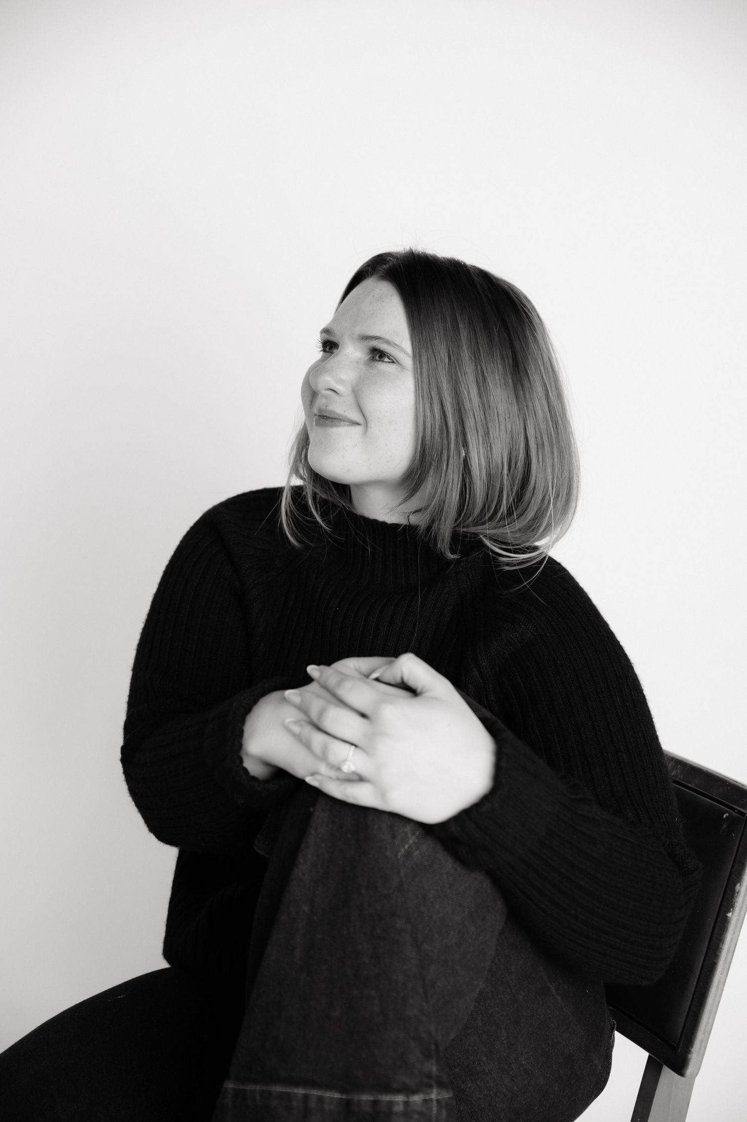 Black and white portrait of a woman in a dark sweater sitting in a chair with hands clasped, looking away.
