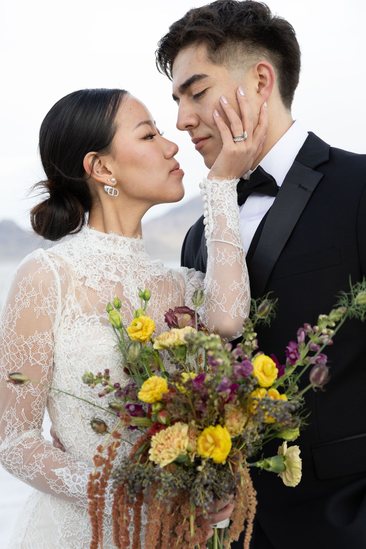 Wedding couple embraces while holding colorful wildflower bouquet against white backdrop.