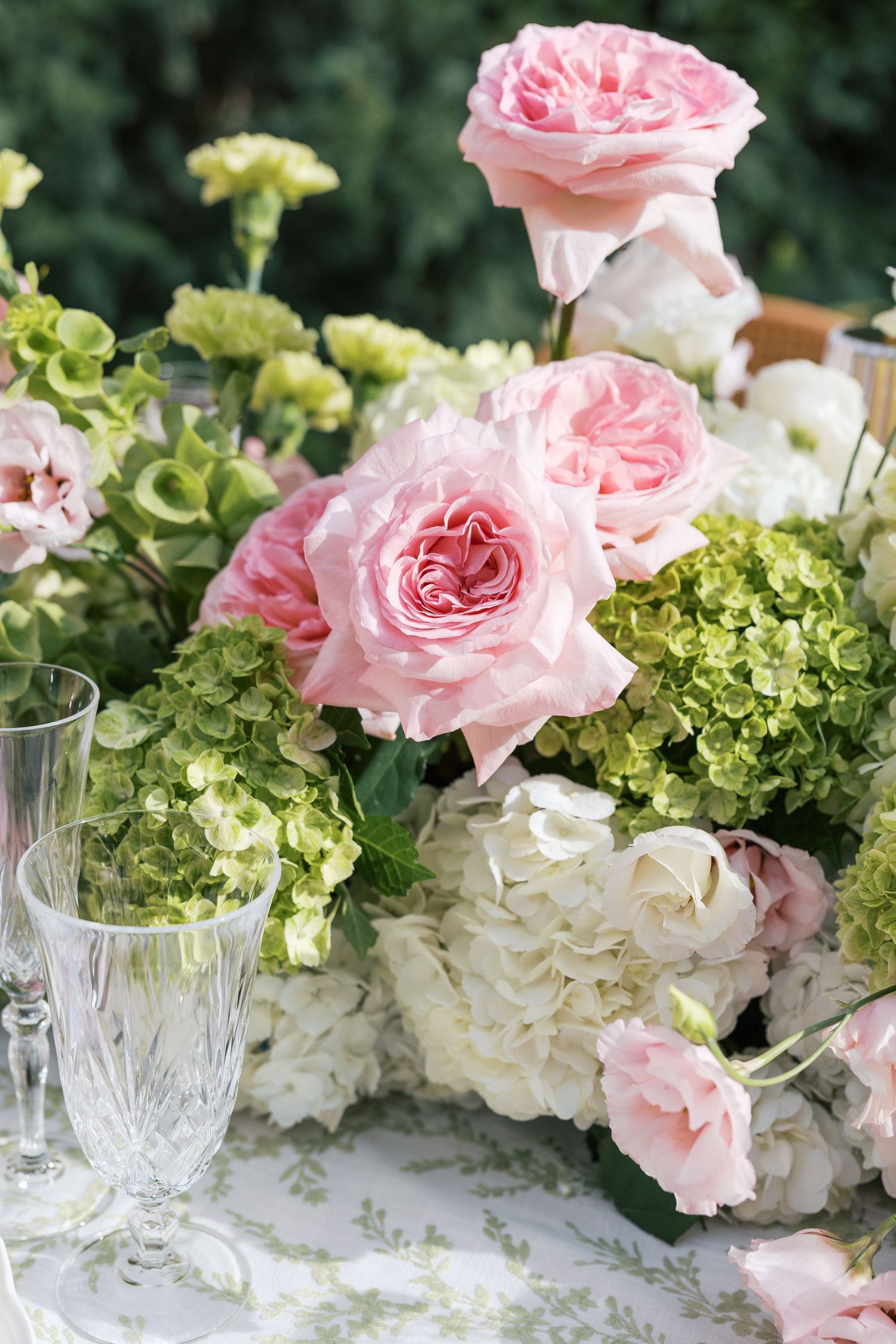 Elegant floral arrangement with pink roses, white hydrangeas, and green blooms beside crystal glasses at an outdoor event.