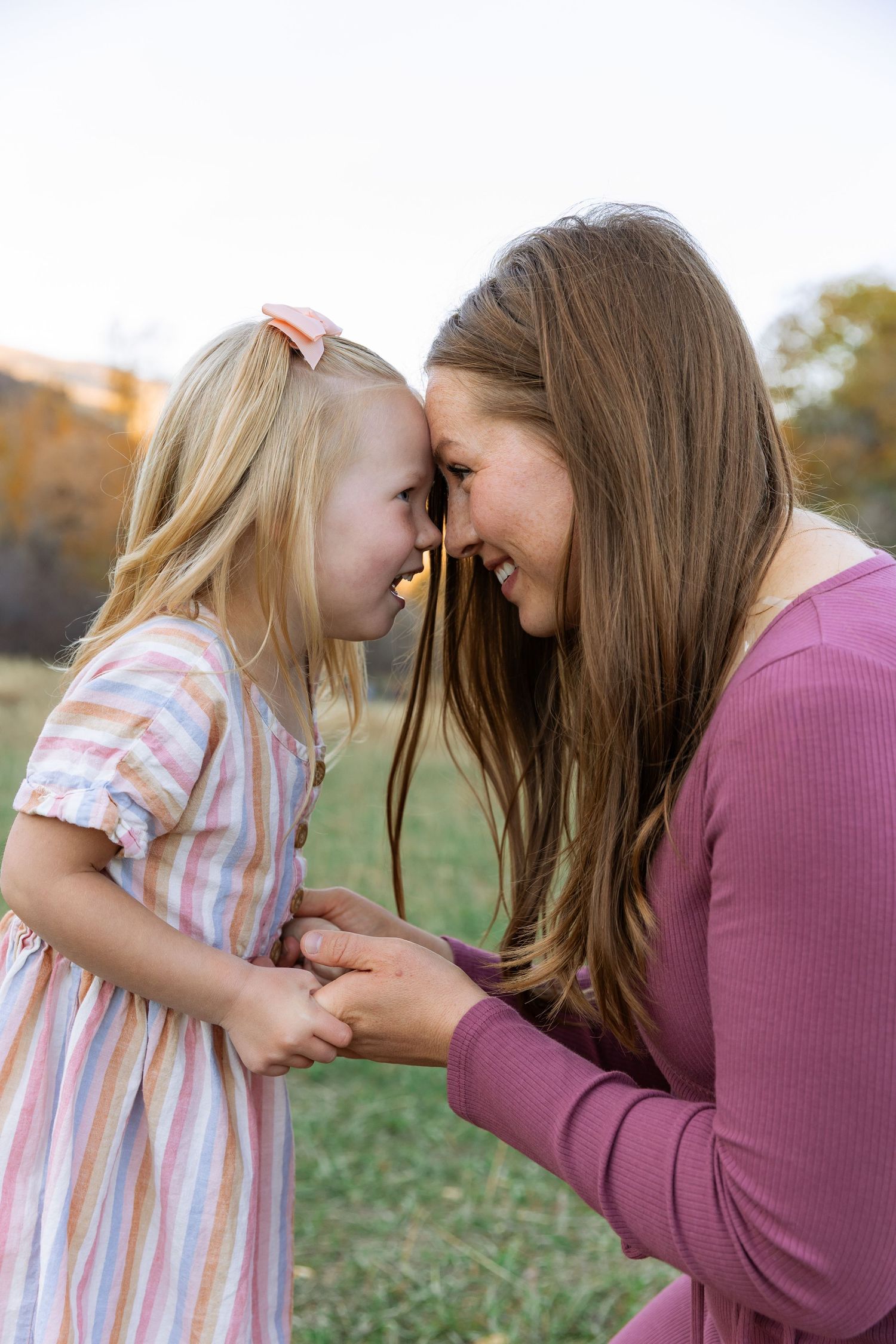 A mother and young blonde girl touch foreheads and smile while holding hands outdoors in a sunny field.
