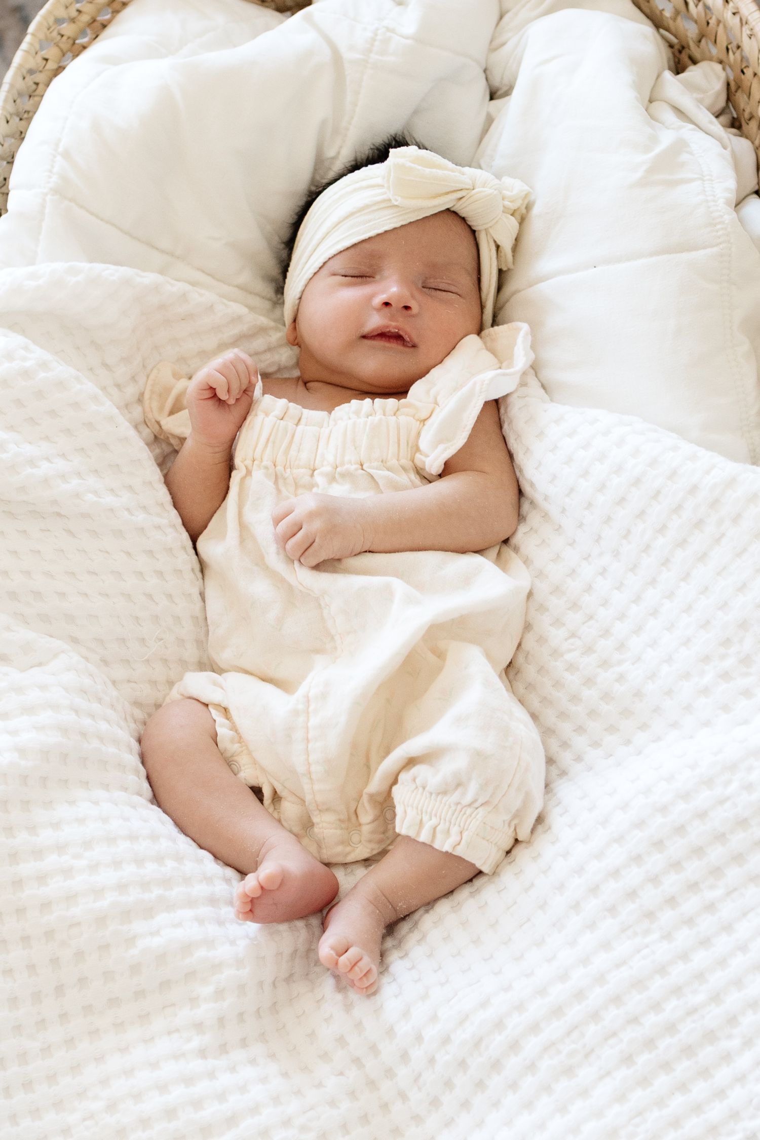 Sleeping newborn baby in cream romper and bow headband, nestled in a white wicker bassinet with soft blankets.