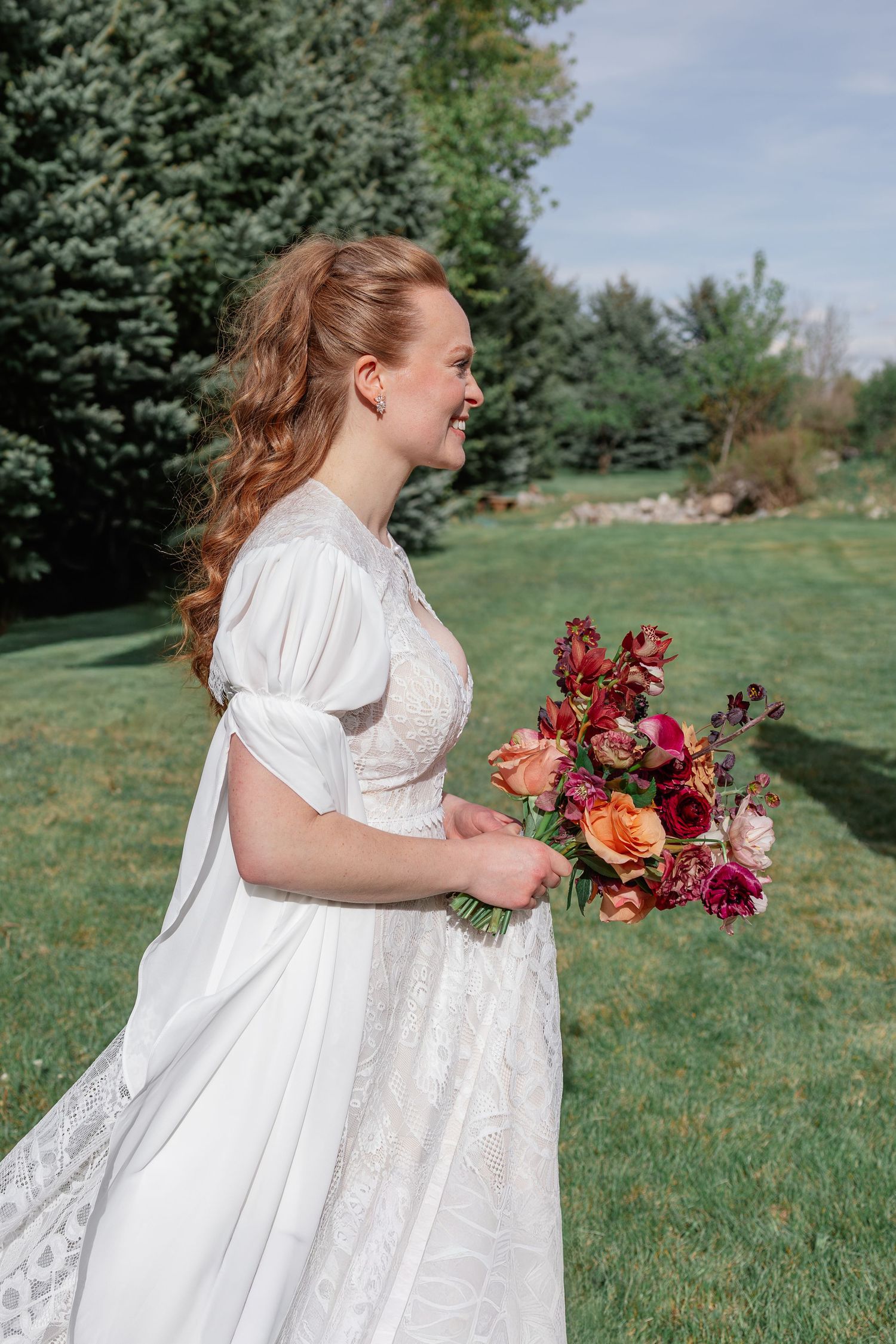 Bride in white lace dress holding vibrant red and orange floral bouquet standing on green lawn.