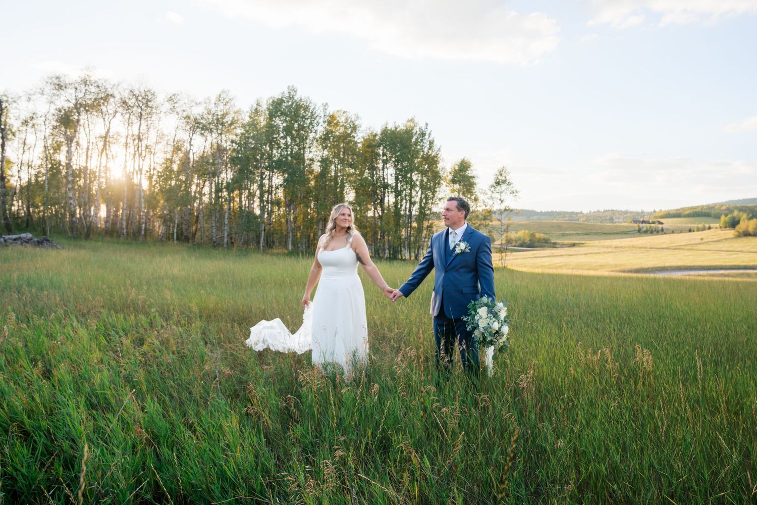 Bride and groom holding hands walking through lush green meadow at golden sunset with tall trees in background.