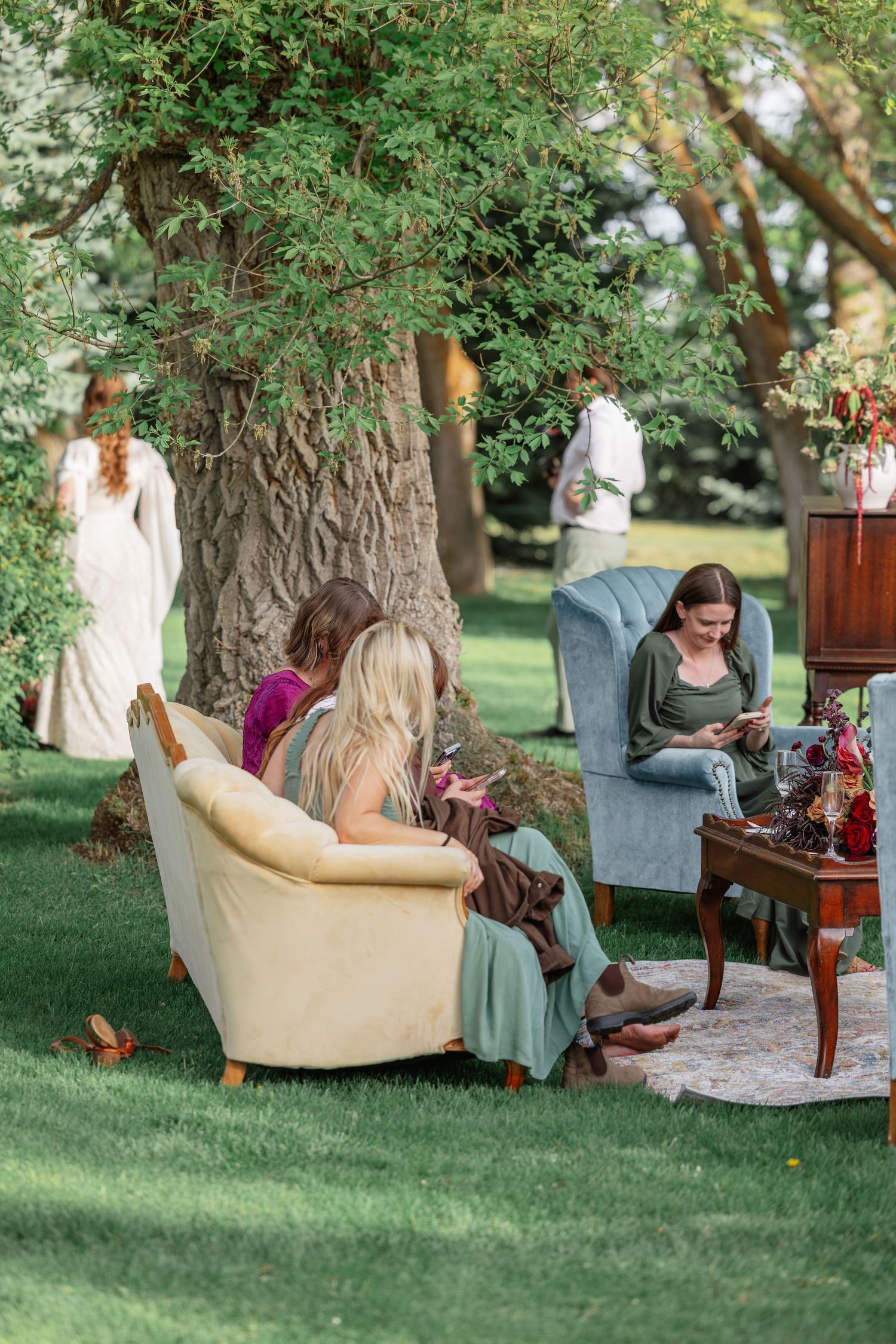 Women relaxing in vintage armchairs on green lawn under large tree at elegant outdoor garden wedding reception.