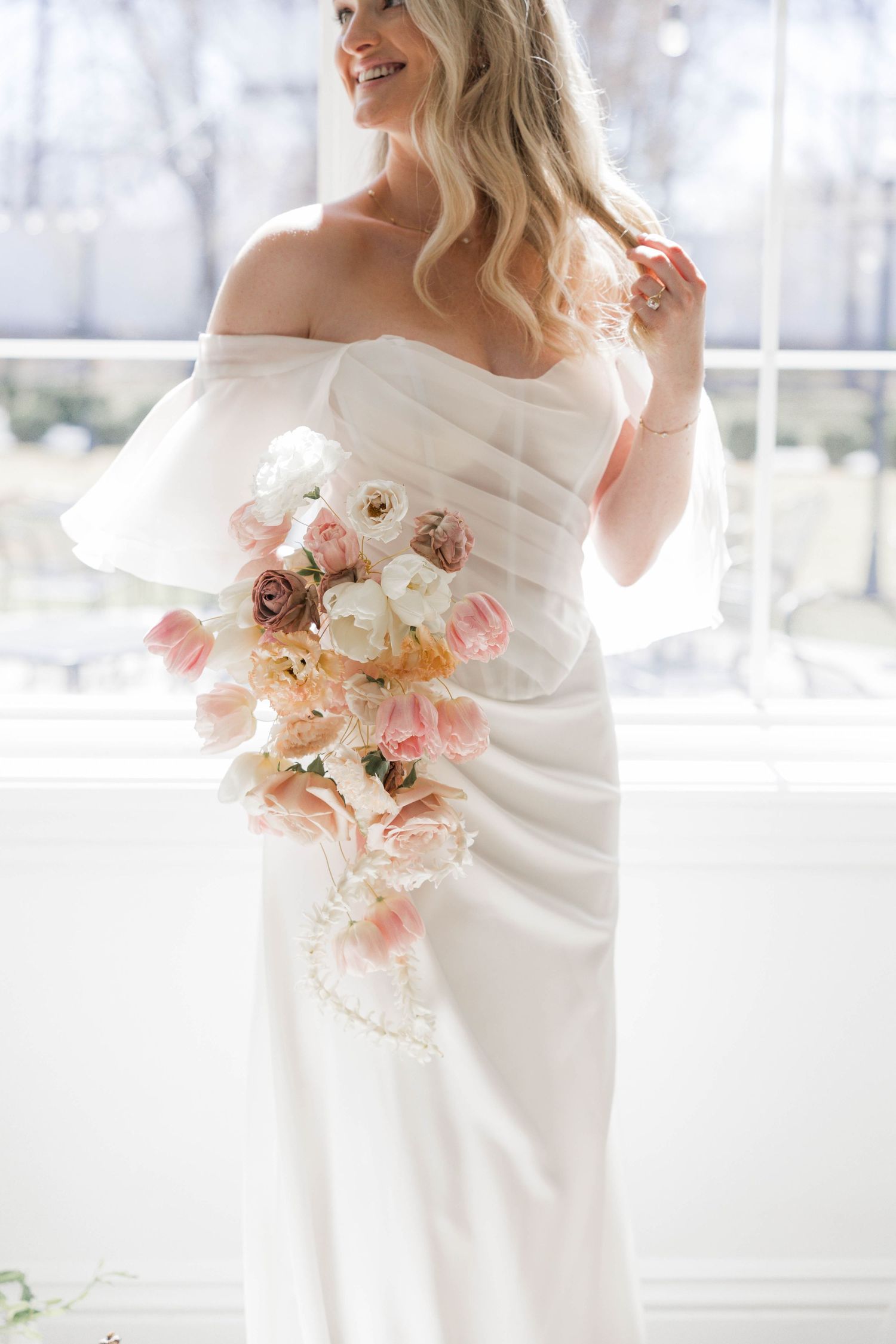 Elegant bride in off-shoulder white gown holds romantic pastel bouquet of pink and cream flowers against bright window.