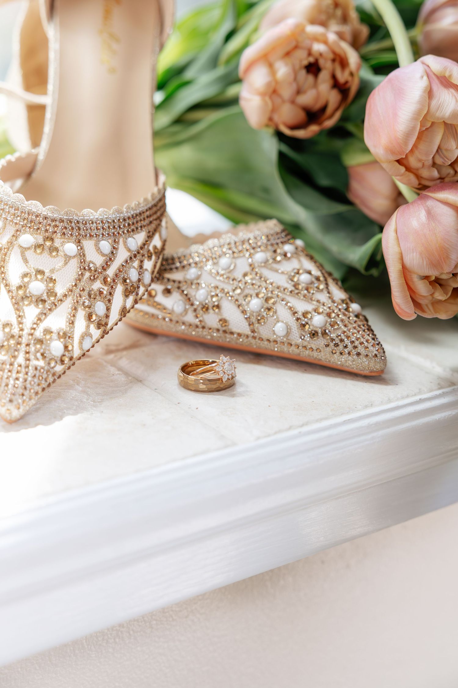 Elegant beaded bridal heels with pearl embellishments beside gold wedding rings and pink tulips.