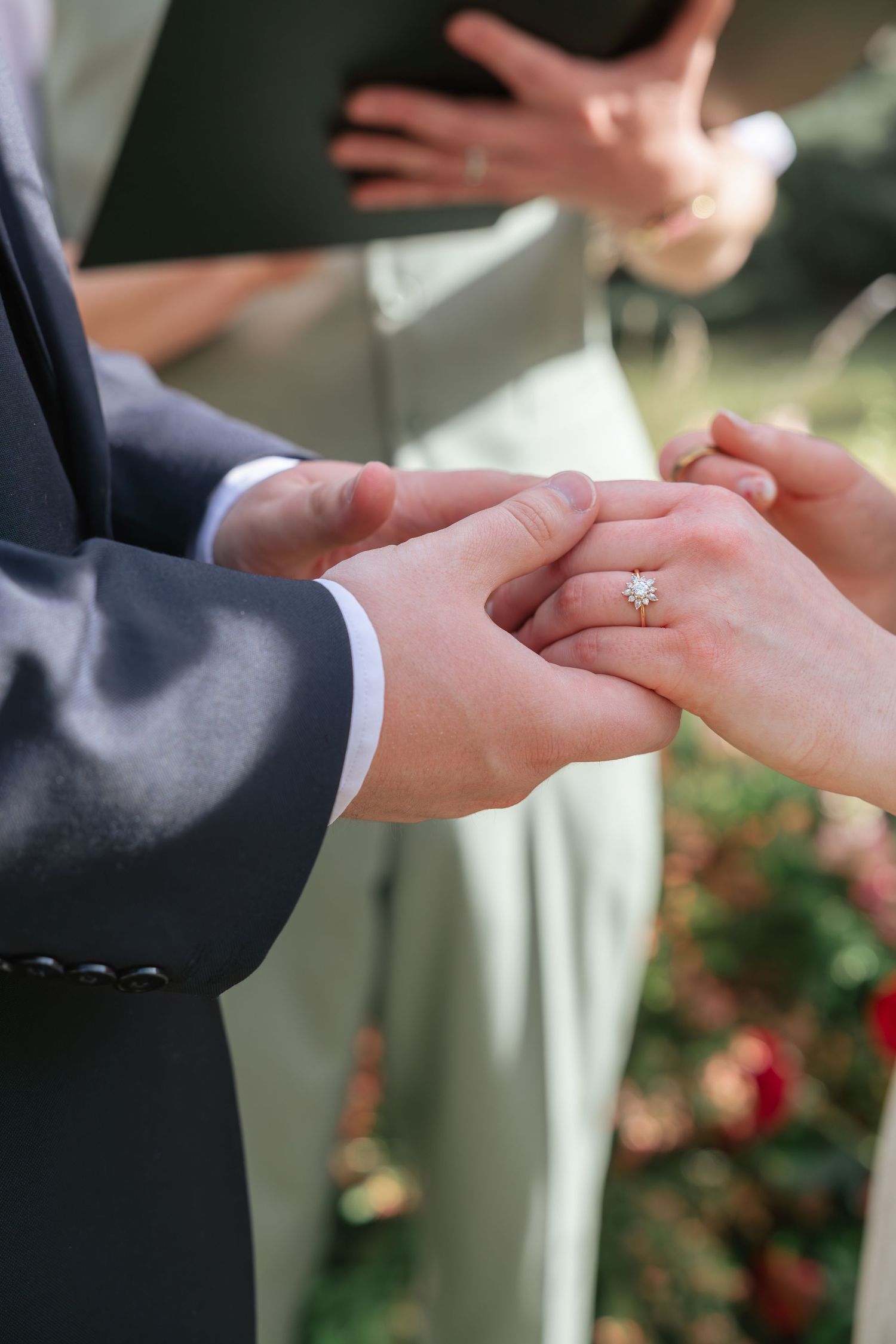 Groom placing a diamond engagement ring on bride's finger during outdoor wedding ceremony with officiant in background.