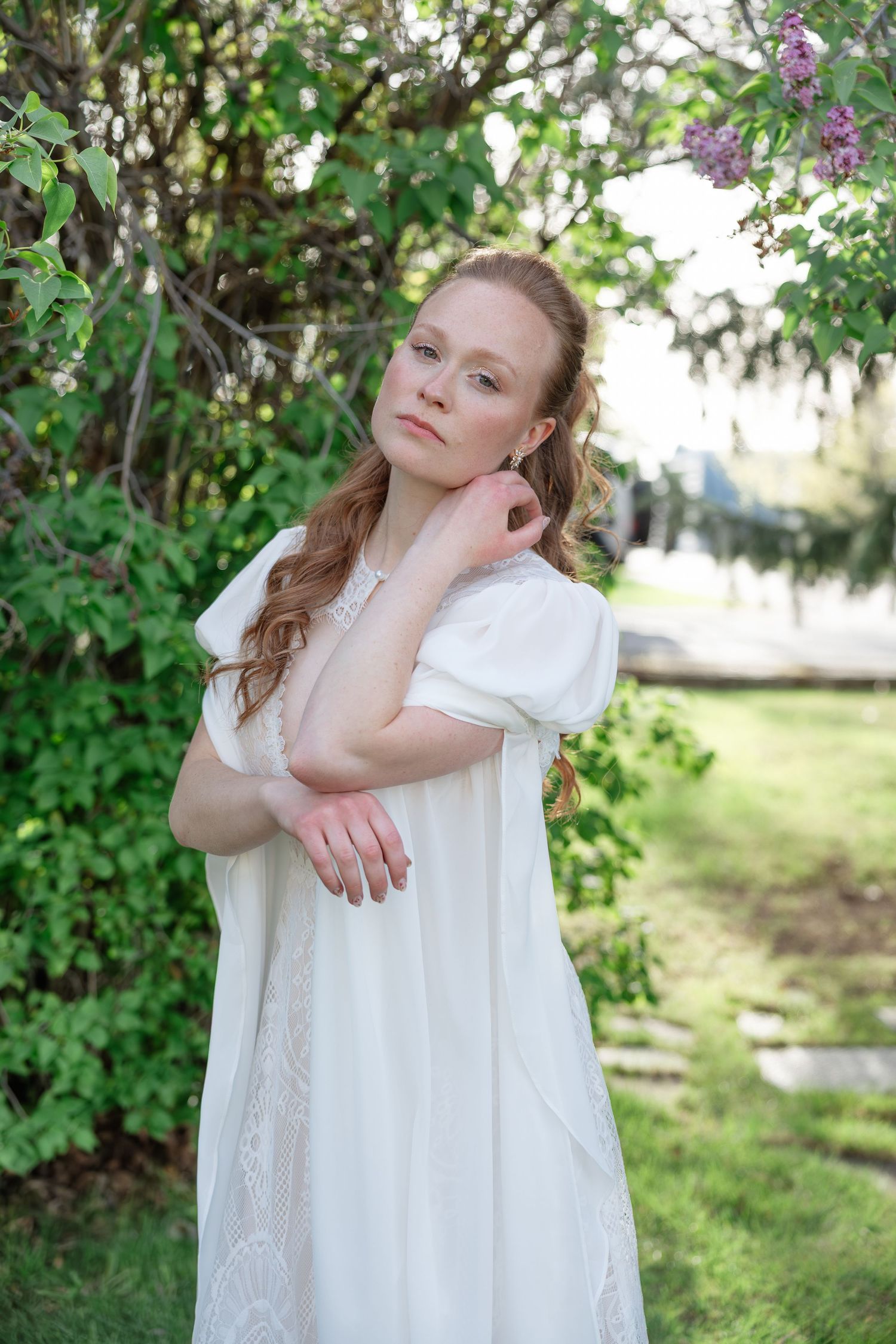 Bride in white lace dress poses gracefully among green foliage and purple lilac blooms outdoors.