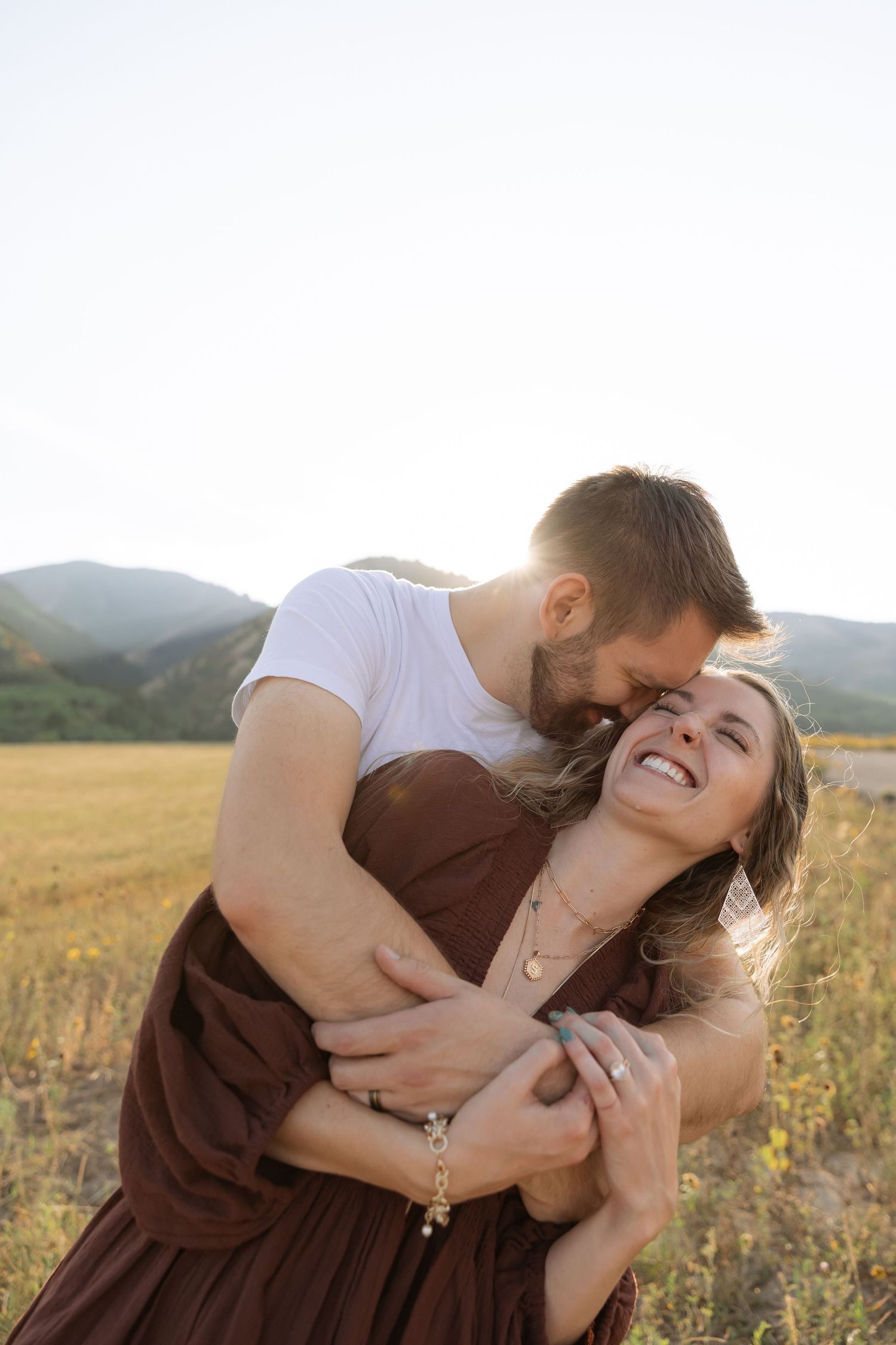 Couple embracing in a golden field with mountains, man kissing woman's head as she laughs joyfully.