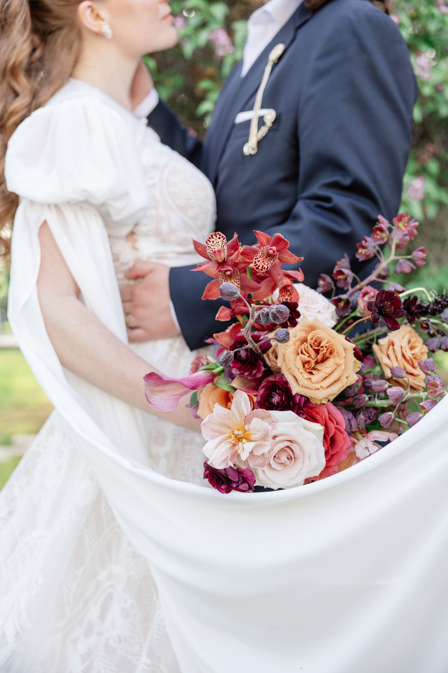 Bride in white lace gown holds stunning bouquet of burgundy, peach, and pink roses with groom.