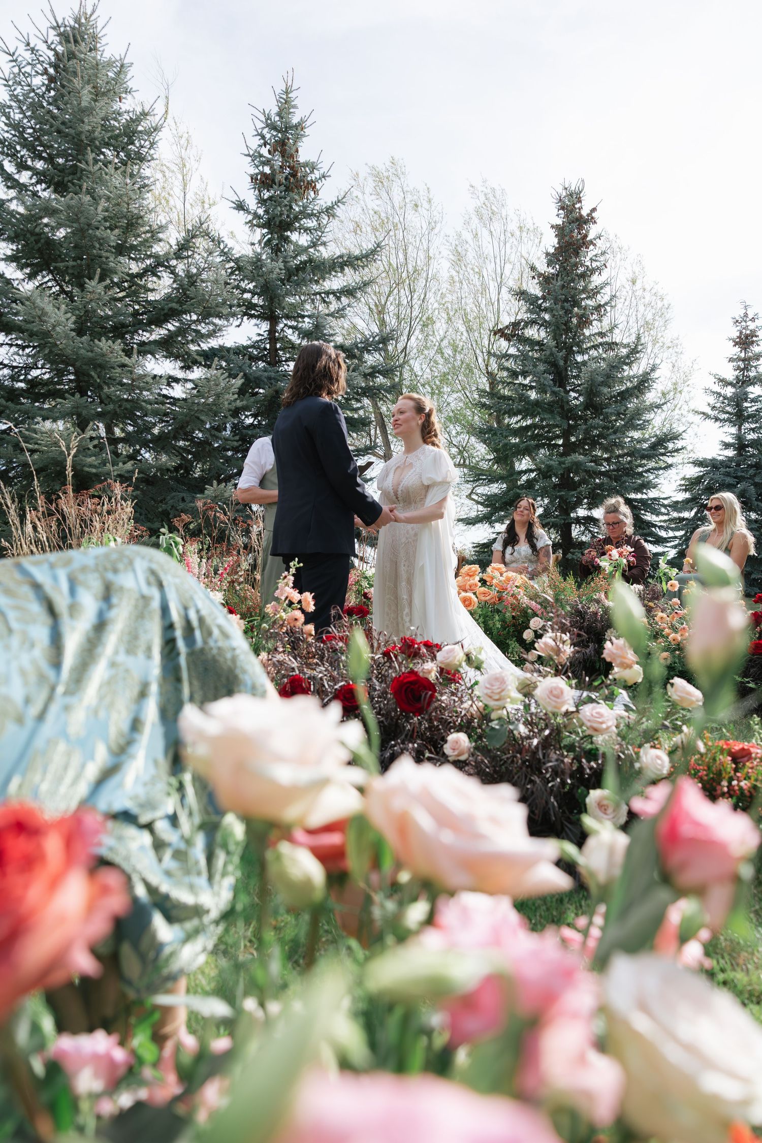 Romantic outdoor wedding ceremony framed by colorful garden flowers and tall pine trees.