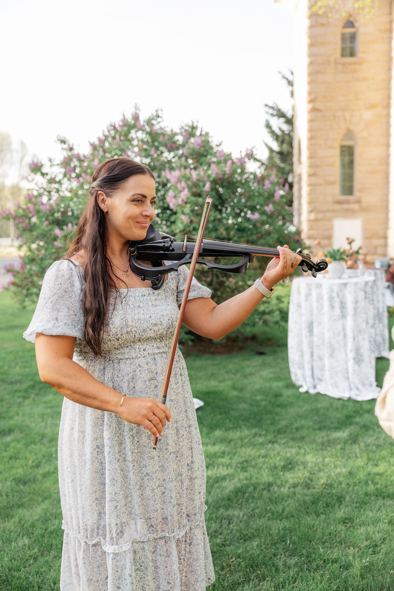 Smiling woman in white floral dress playing black violin outdoors near blooming lilac bushes and stone church tower.