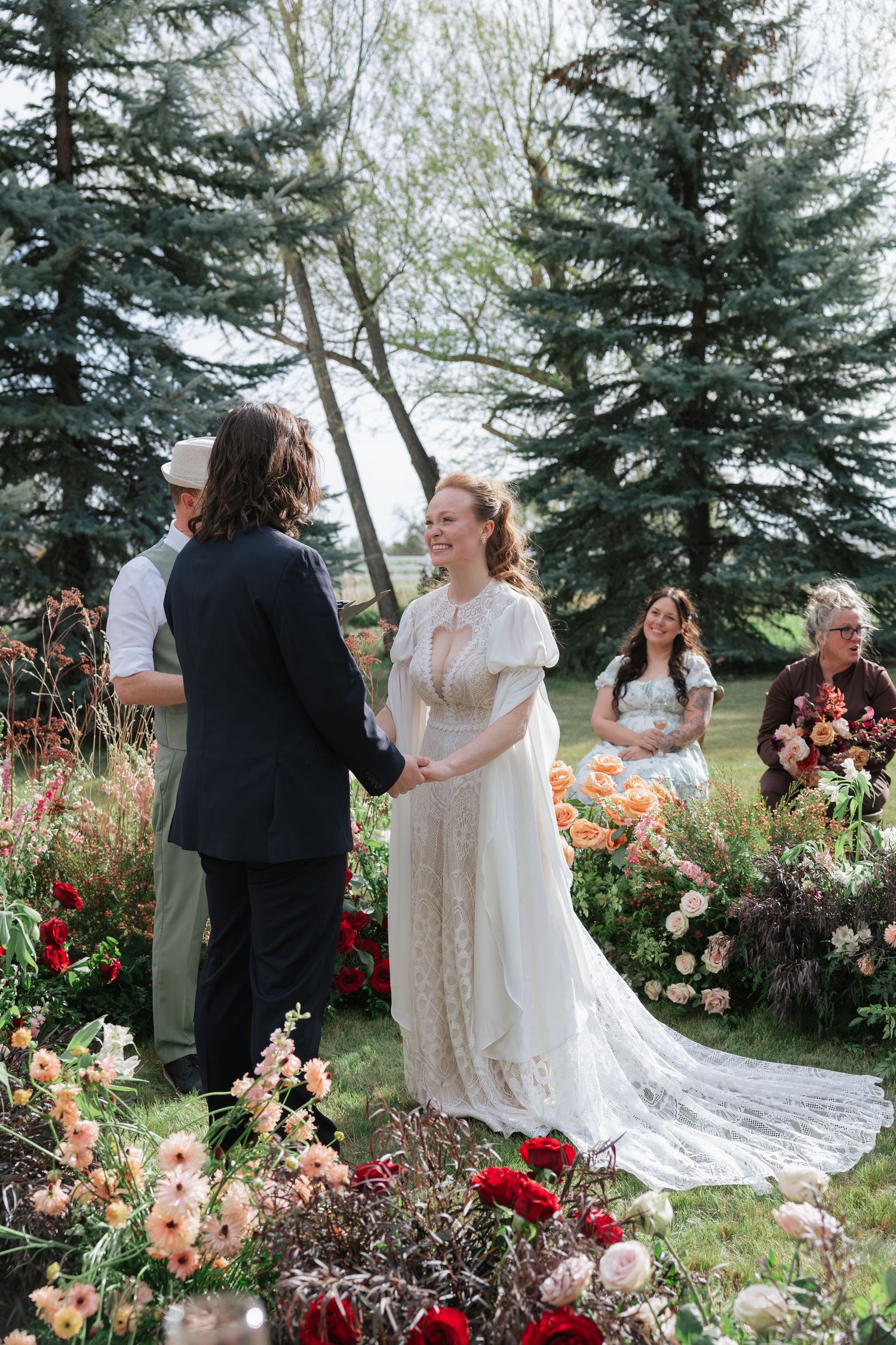 Bride and groom exchange vows outdoors surrounded by lush garden florals and evergreen trees.