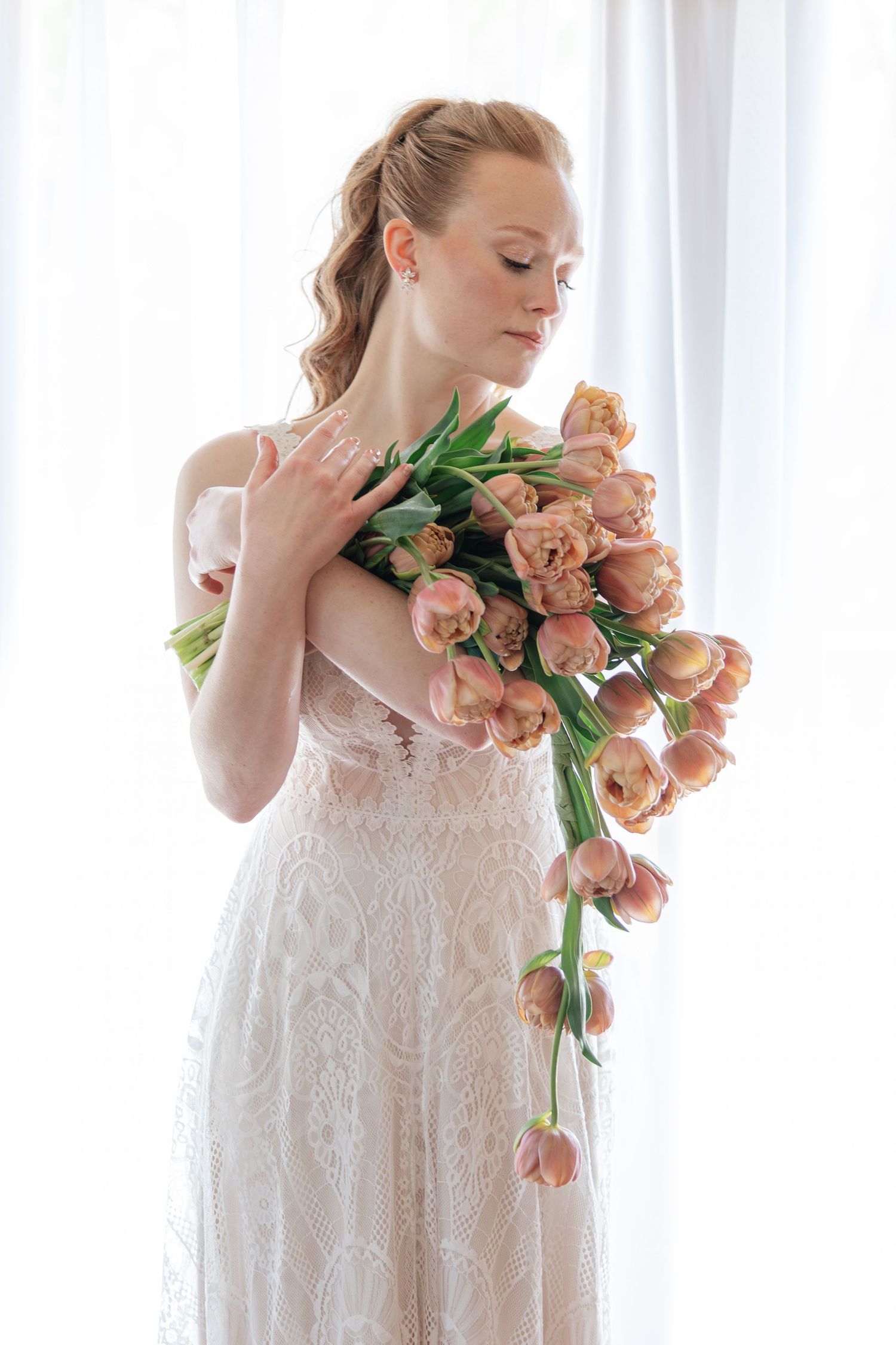 Elegant bride in lace dress holding large bouquet of pink tulips near white curtains. Shelton Inn Idaho Falls