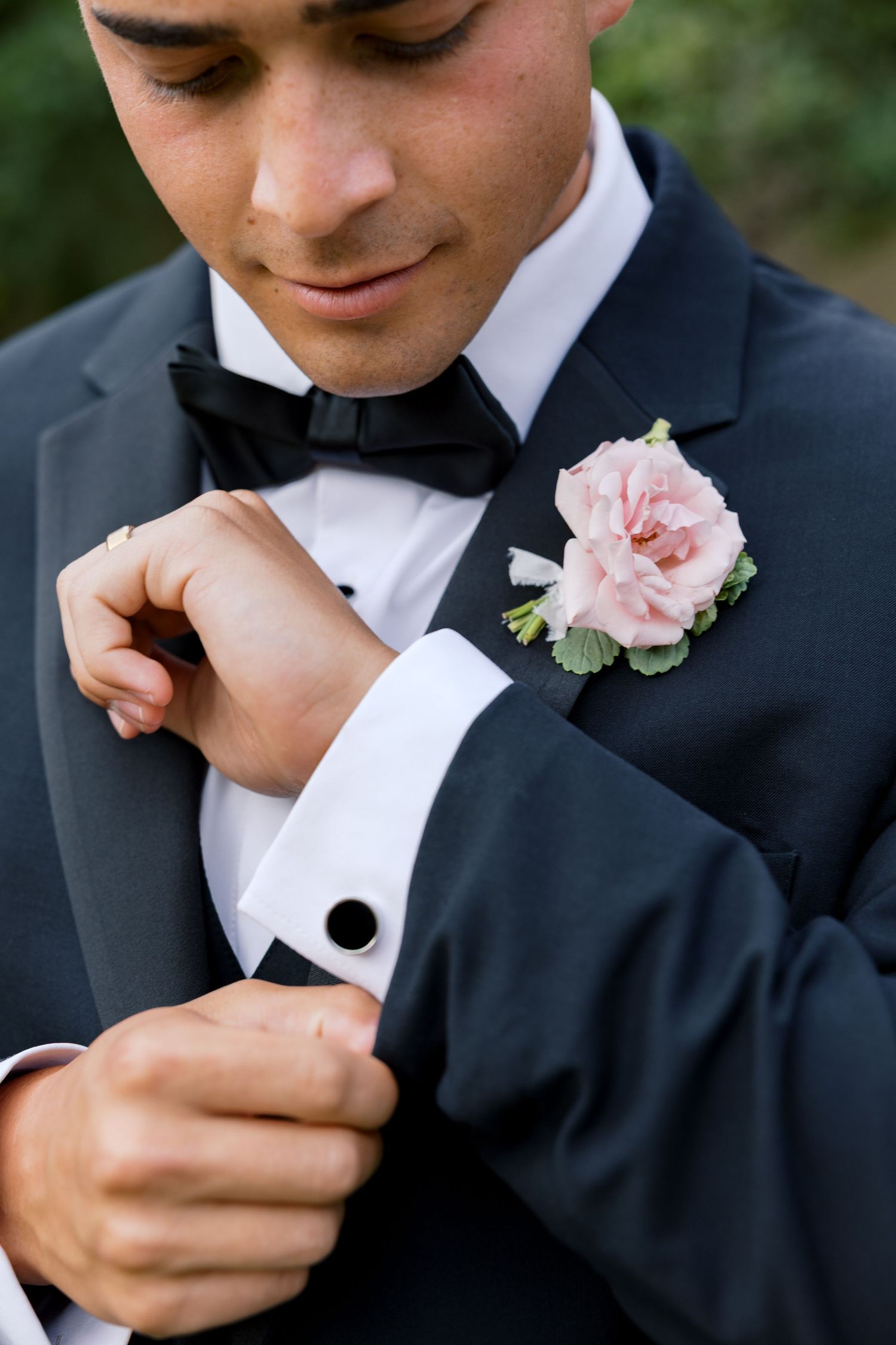 Groom in black tuxedo with bow tie adjusting cufflinks, wearing a pink rose boutonniere on his lapel.