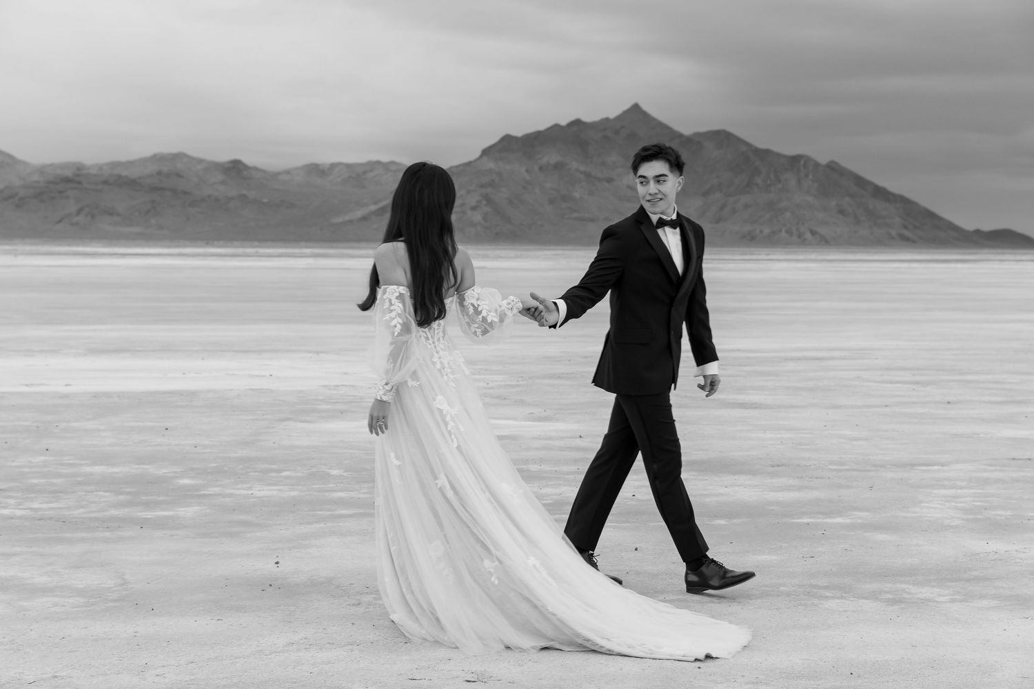 A black and white photo of newlyweds dancing on salt flats with mountains in the distance.