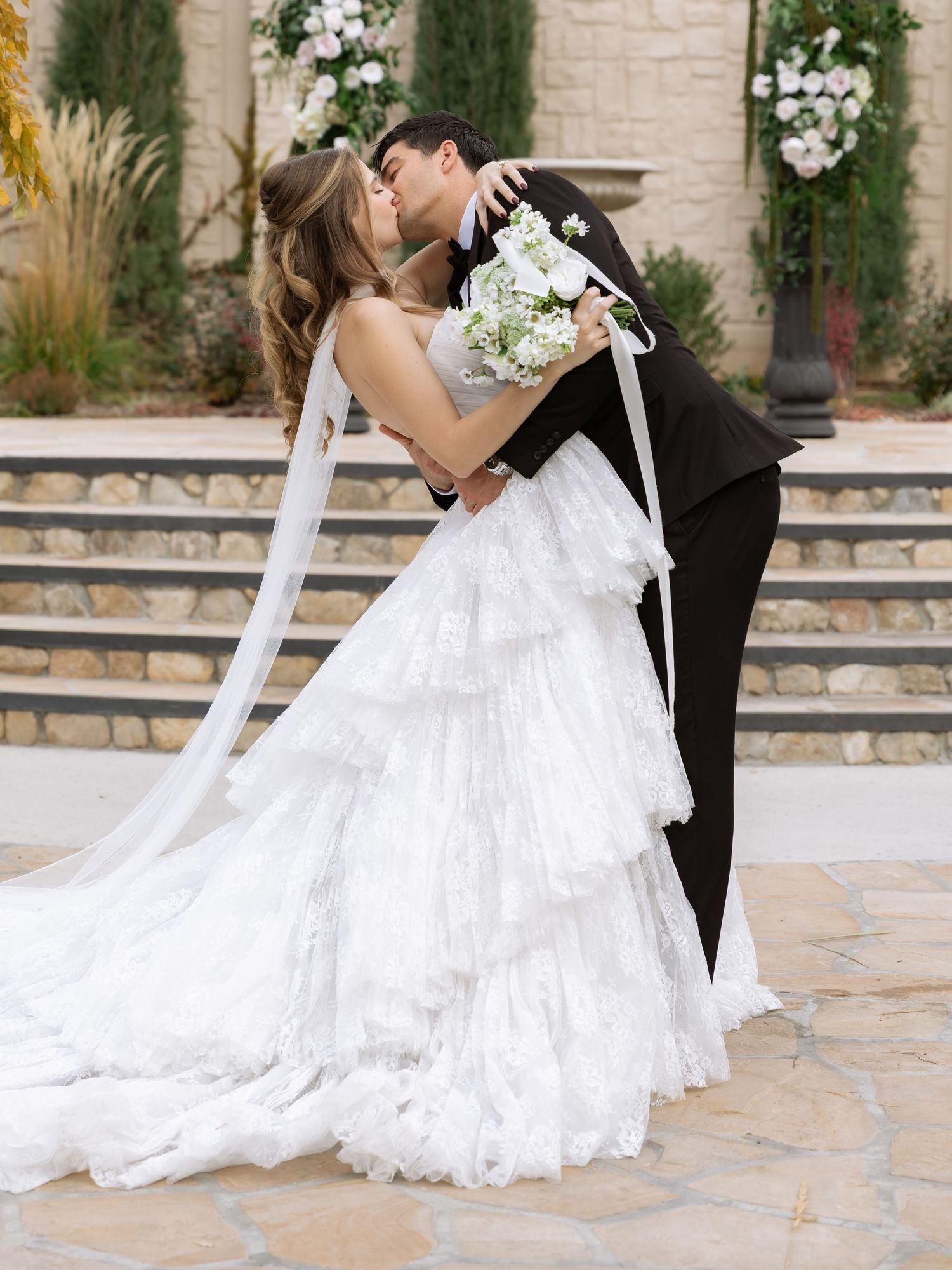 Romantic wedding couple shares intimate moment on stone patio with floral backdrop.
