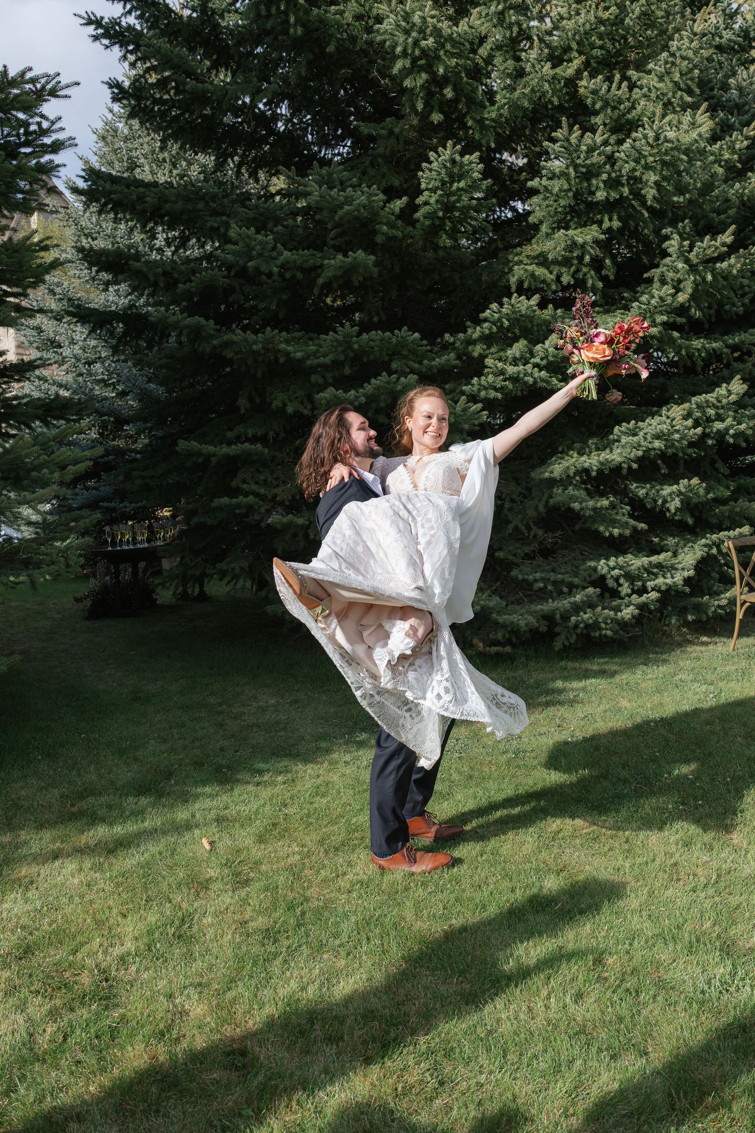 Joyful groom lifting bride in white lace gown as she raises her bouquet in a sunny outdoor garden setting.
