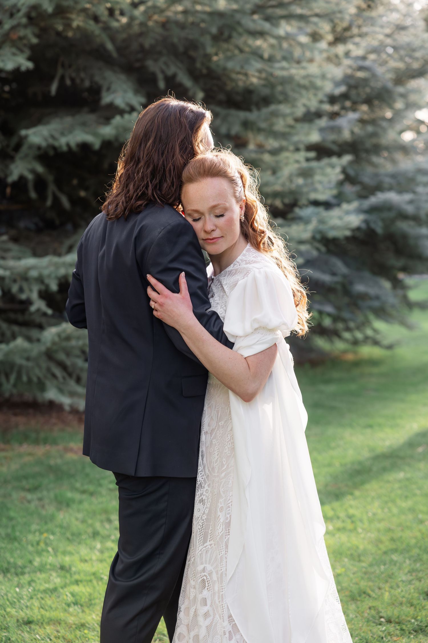 Bride in white lace gown embracing groom in dark suit near tall evergreen trees outdoors.