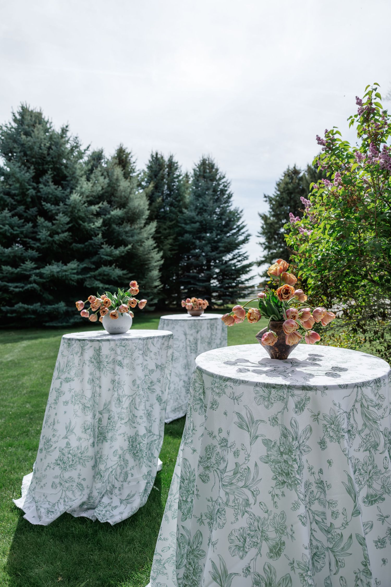 Outdoor cocktail tables with green floral tablecloths and peach flower arrangements on a lawn with evergreen trees.
