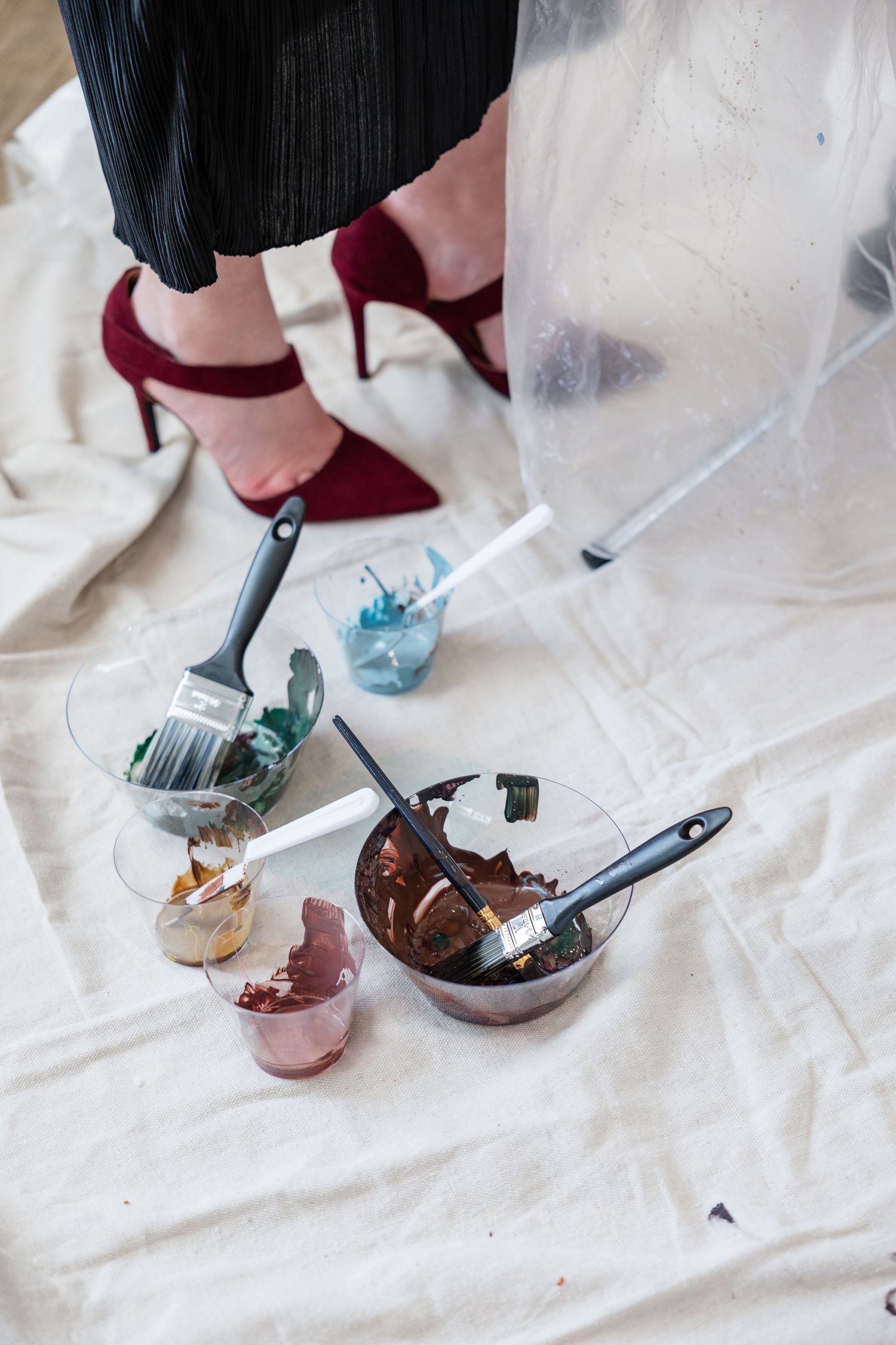 Bowls of hair dye with brushes on a drop cloth, beside a person wearing red high heels.