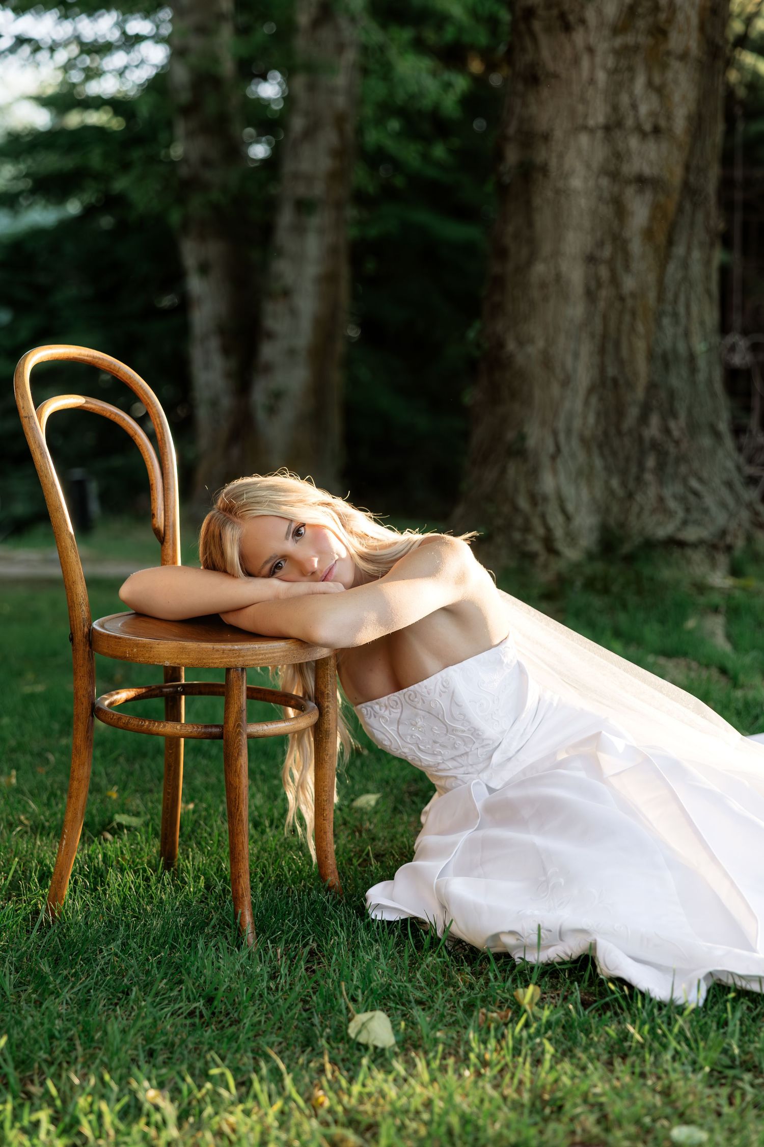 Blonde bride in white gown resting on wooden chair in lush green garden setting.