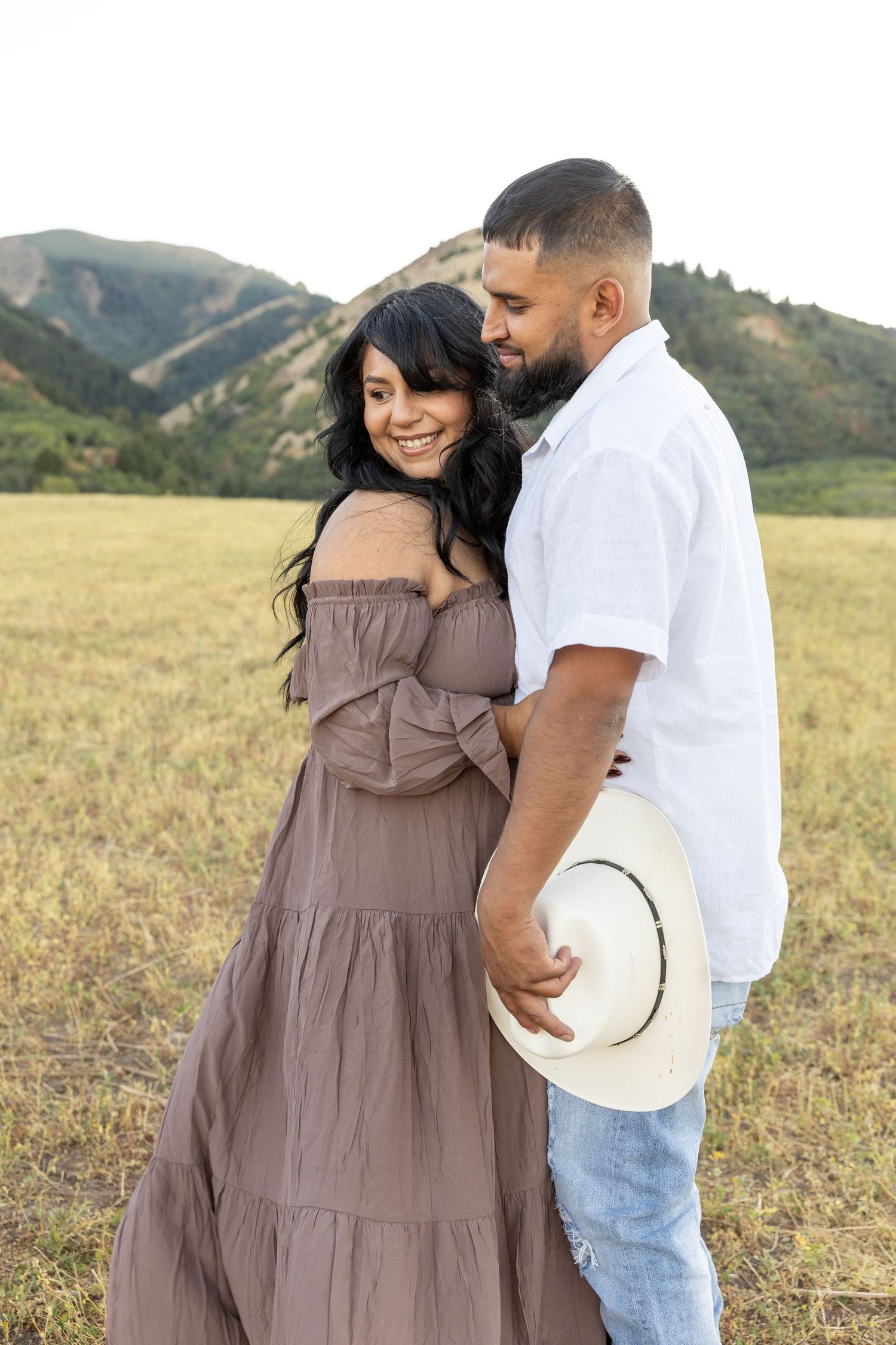 A couple embraces in a mountain meadow, woman in brown dress smiling, man in white shirt holding a cowboy hat.