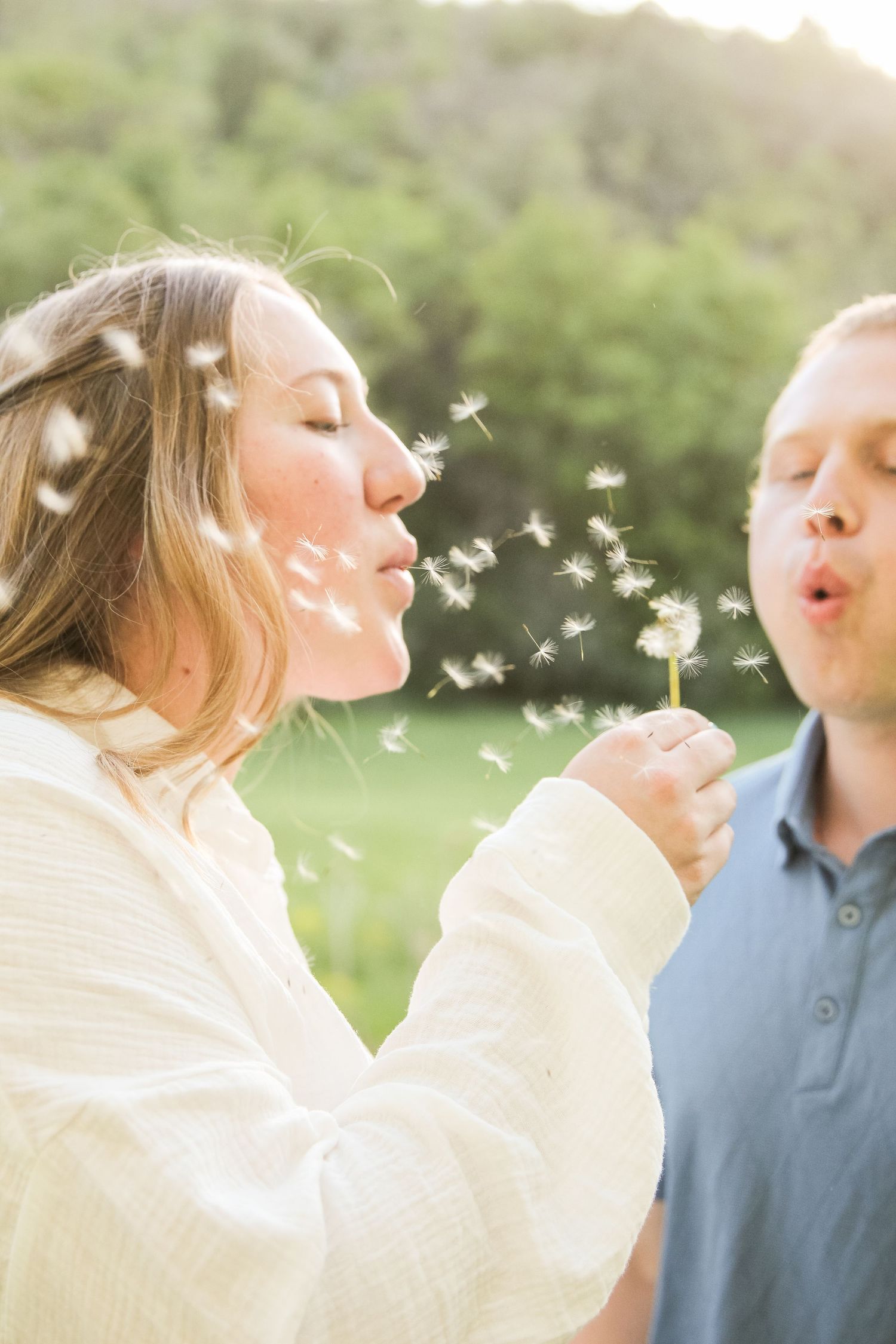 A couple blowing dandelion seeds together in a sunny green field, sharing a romantic outdoor moment.