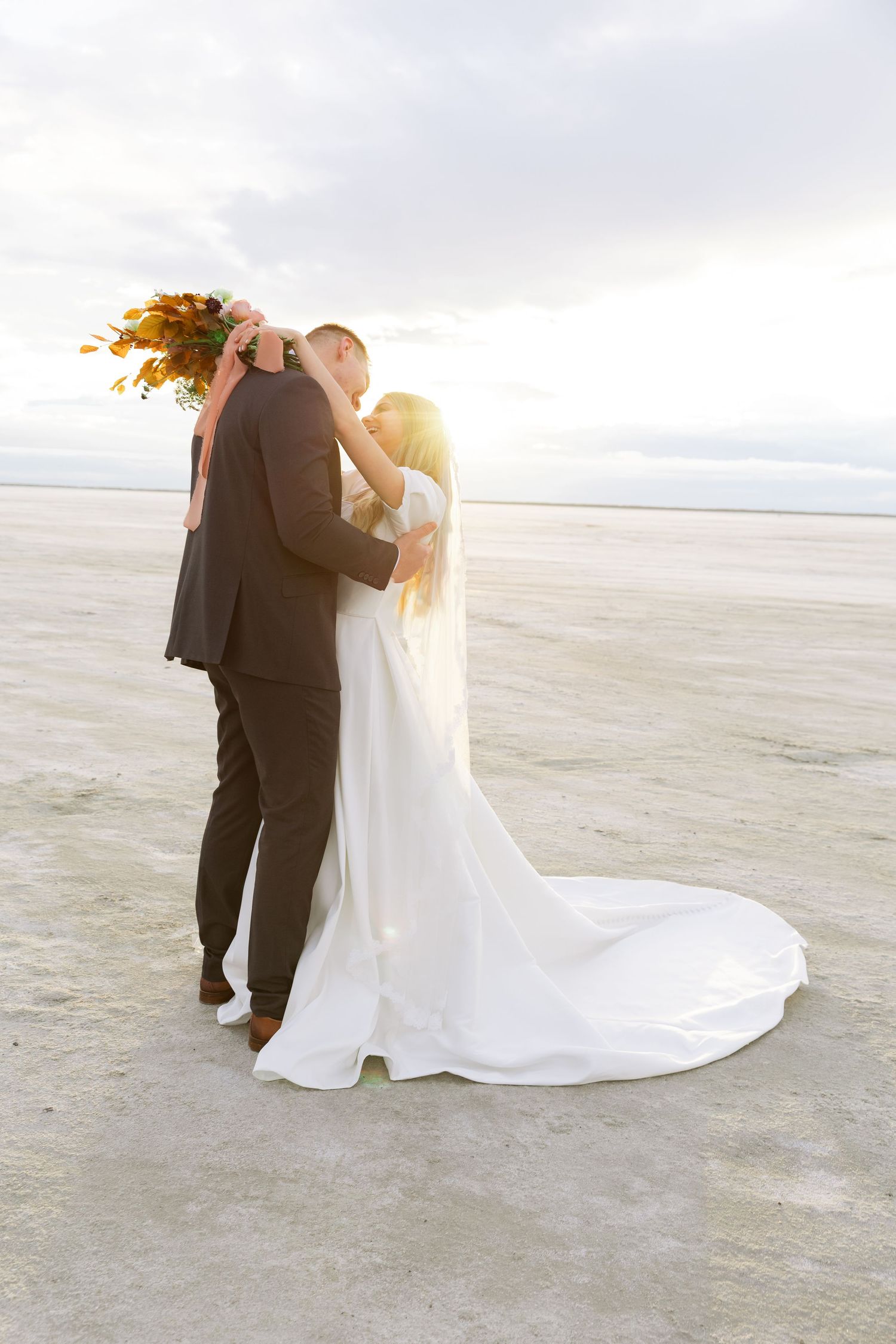 Wedding couple embracing on beach at sunset with flowing white dress.