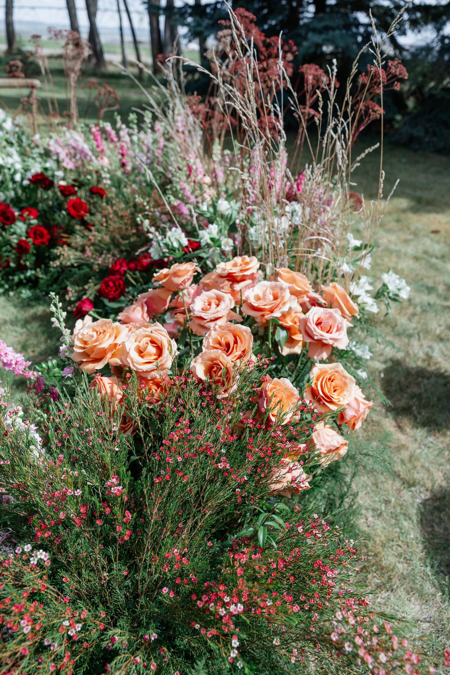 Lush garden floral arrangement featuring peach roses, red blooms, and wildflowers in an outdoor setting.