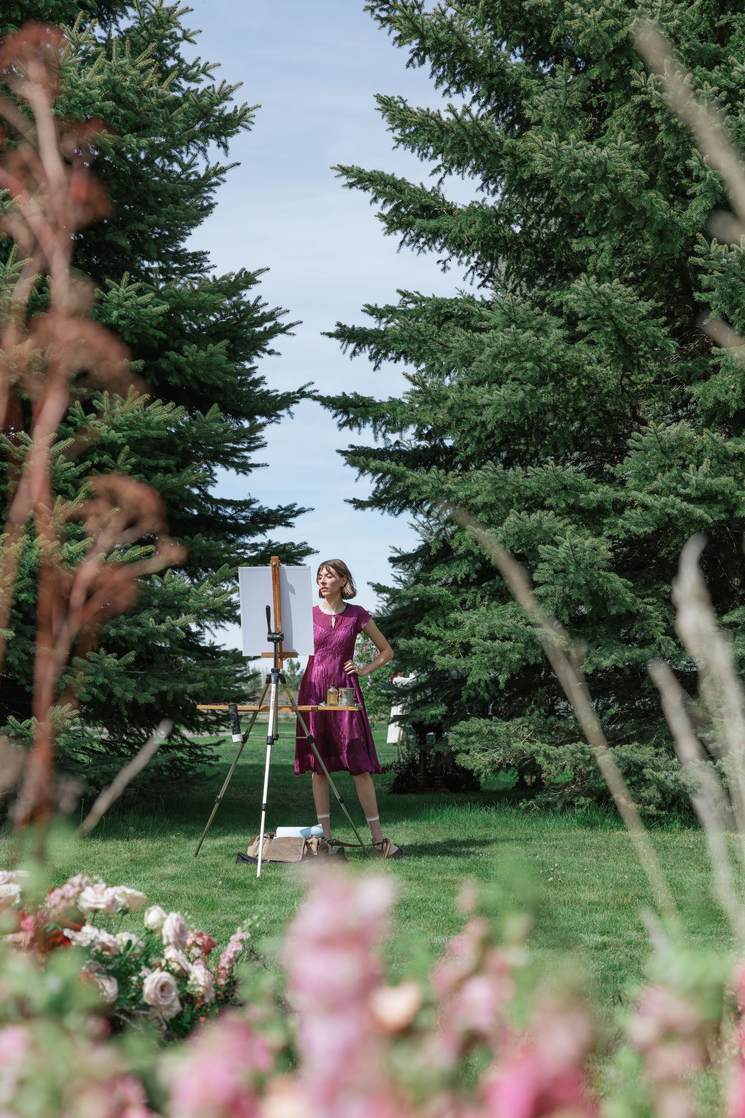 Woman in purple dress painting on easel outdoors surrounded by lush green pine trees and pink flowers.