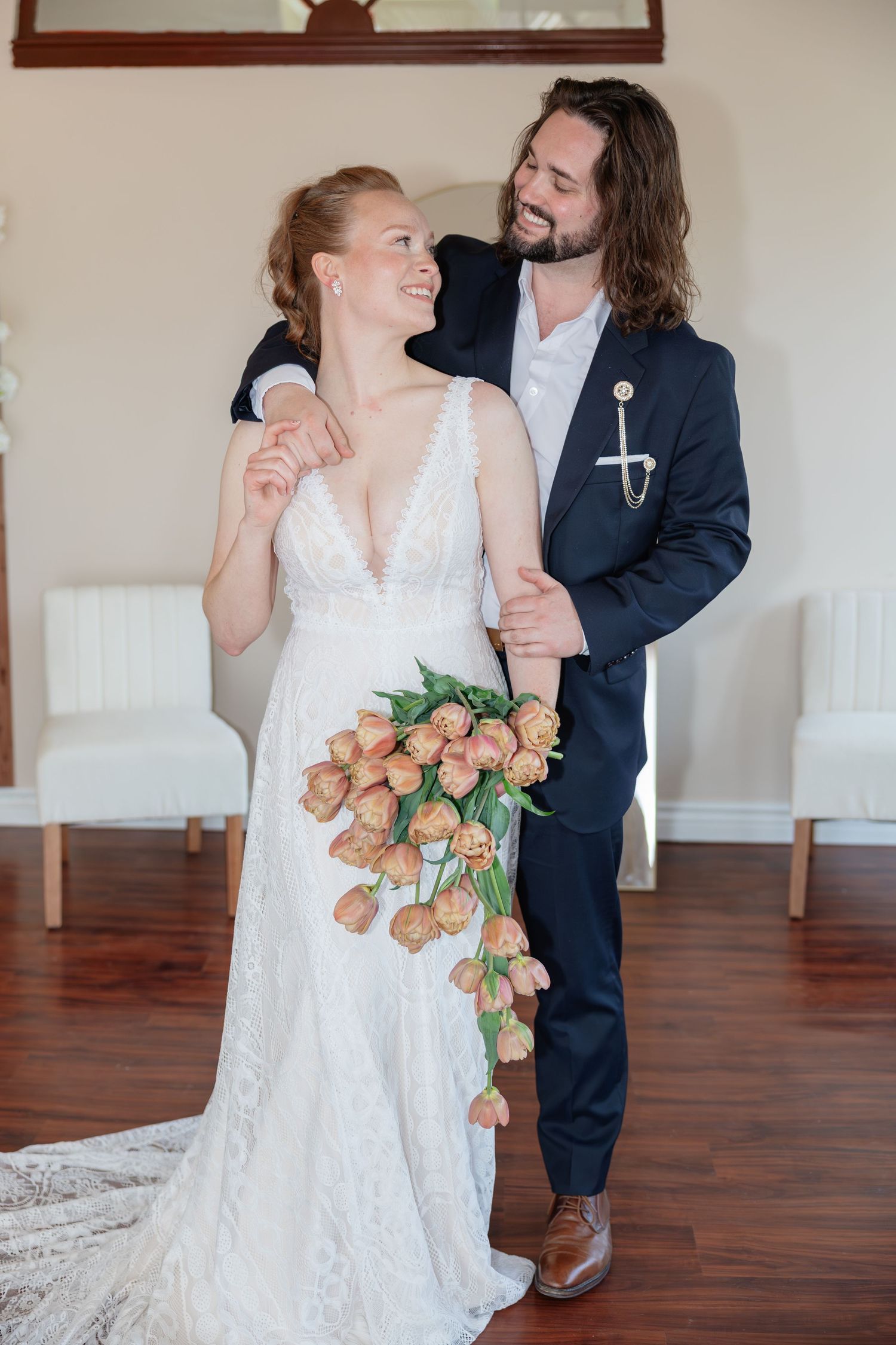 Smiling bride and groom pose indoors, bride holding cascading peach tulip bouquet in lace gown.