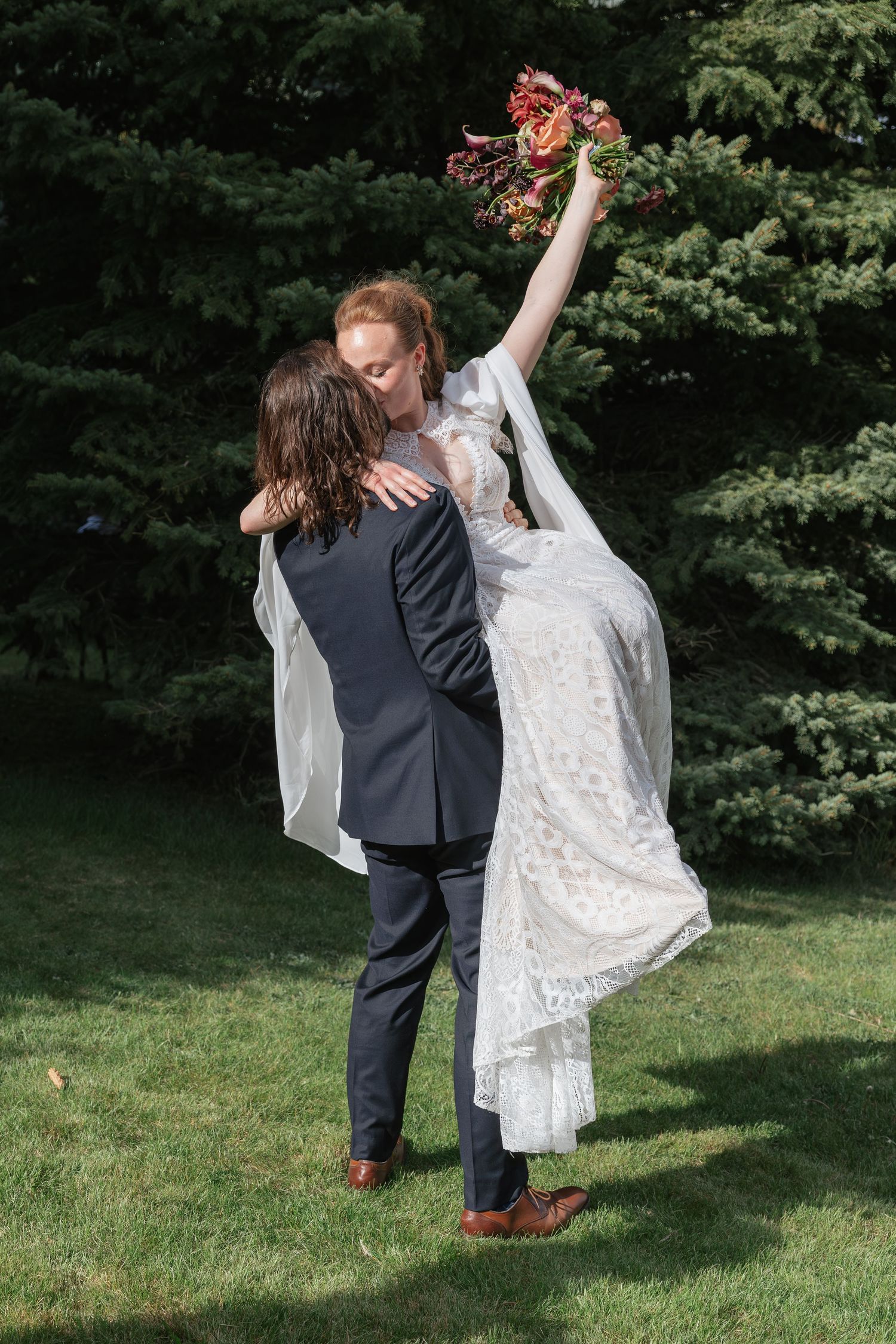 Groom lifting bride in white lace wedding dress as she raises her floral bouquet triumphantly in a lush green garden.