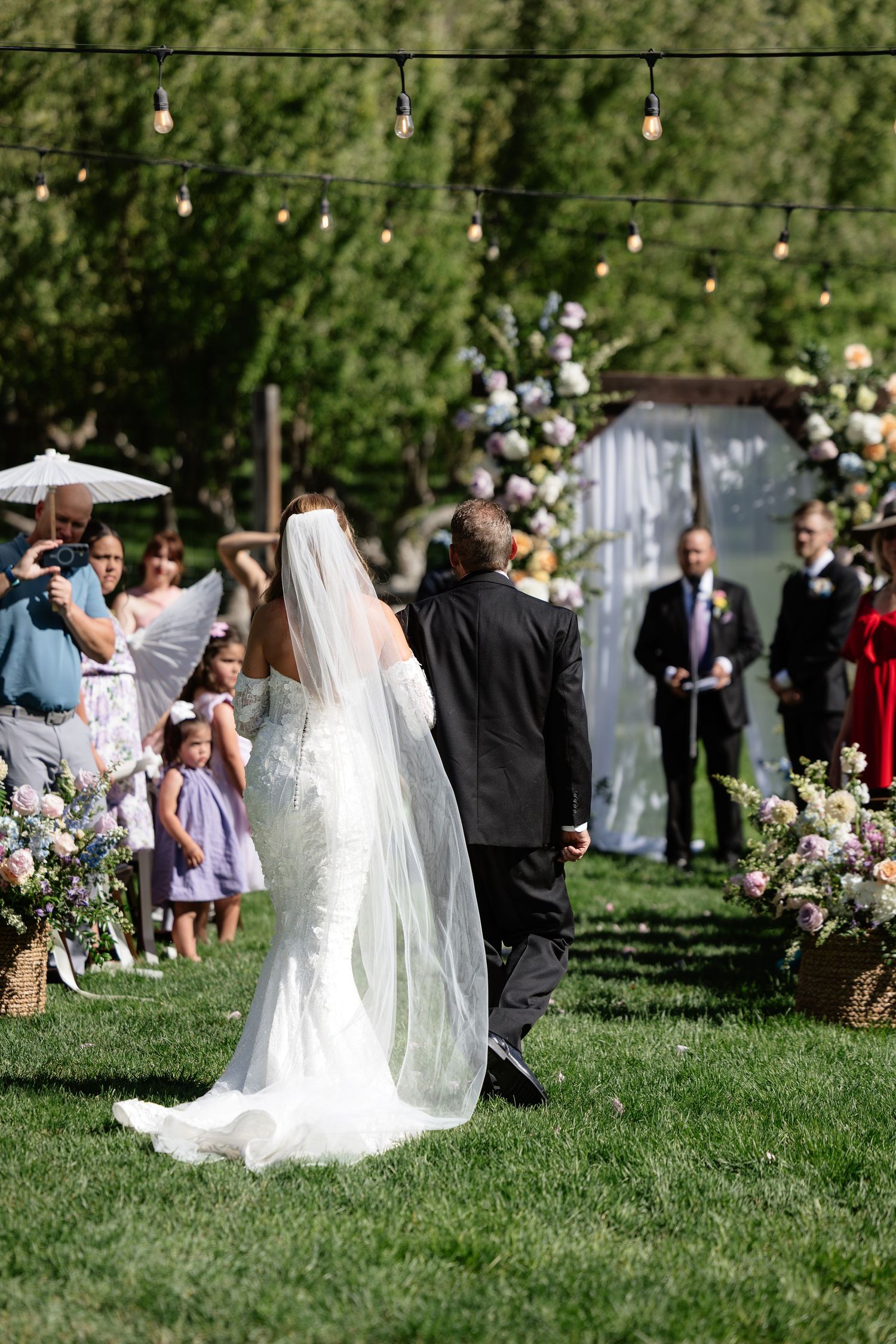 Bride walking down outdoor aisle with escort toward floral arch, guests watching, string lights overhead on sunny wedding day.