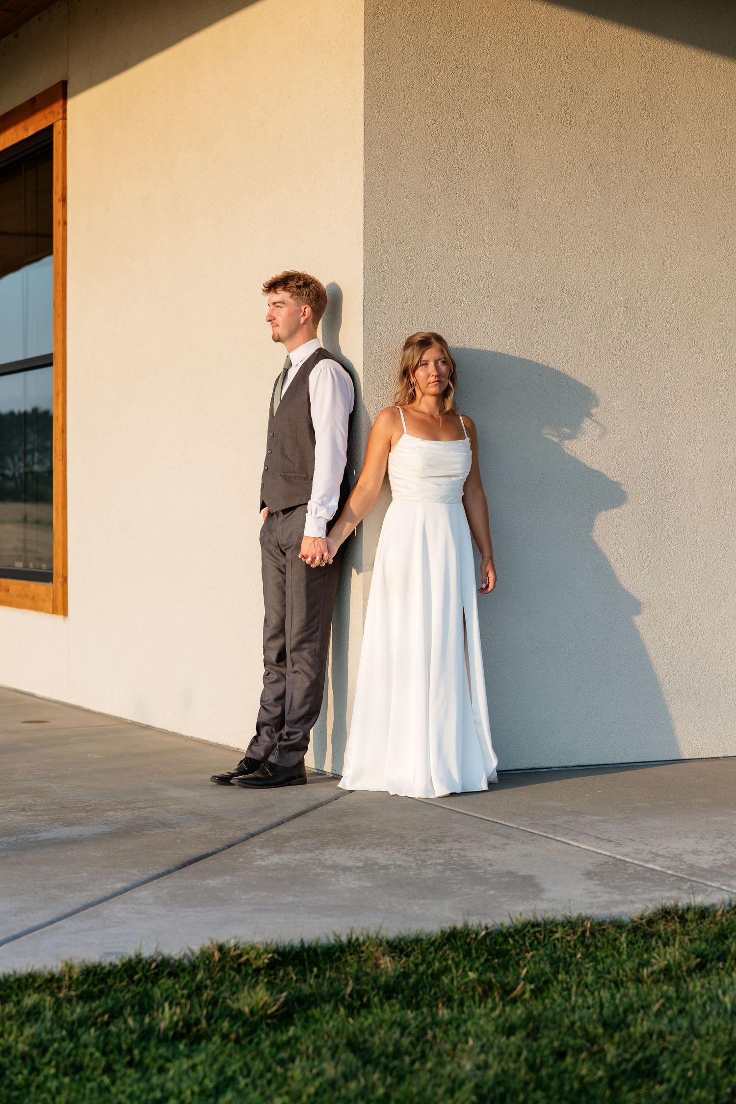 Bride in white gown and groom in gray vest hold hands against a sunlit stucco wall outdoors.