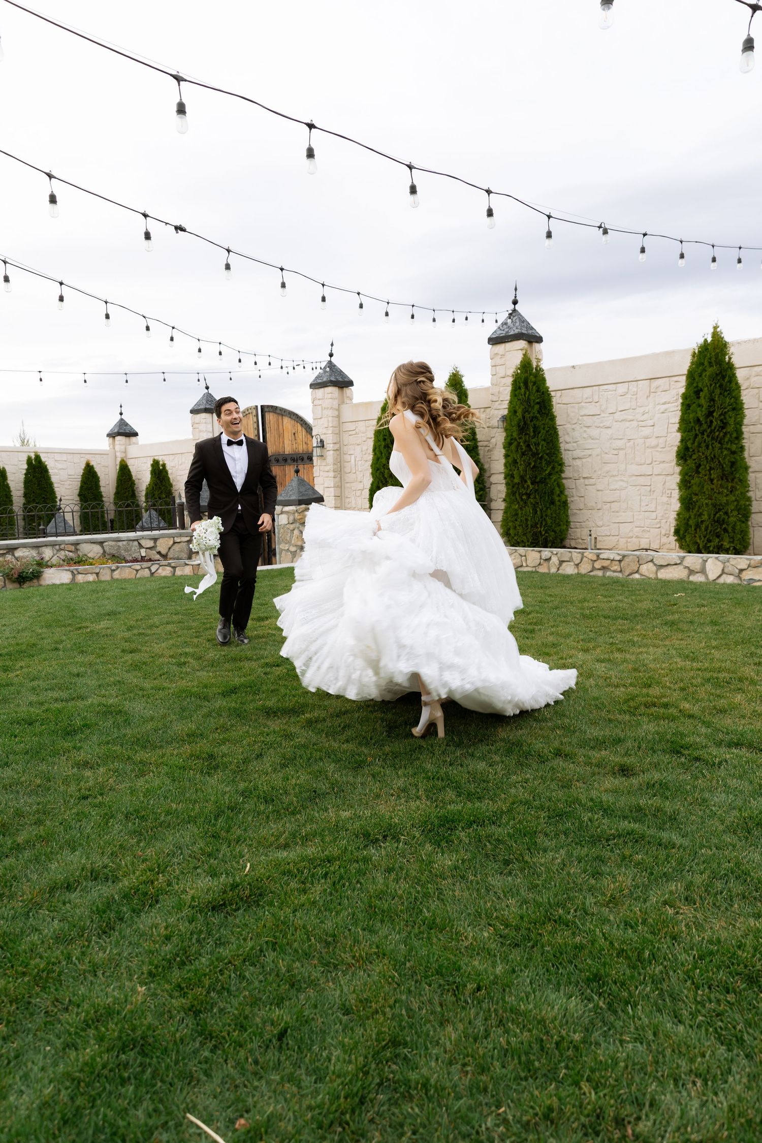 Bride in flowing white gown runs toward groom in black tuxedo on green lawn with string lights overhead.