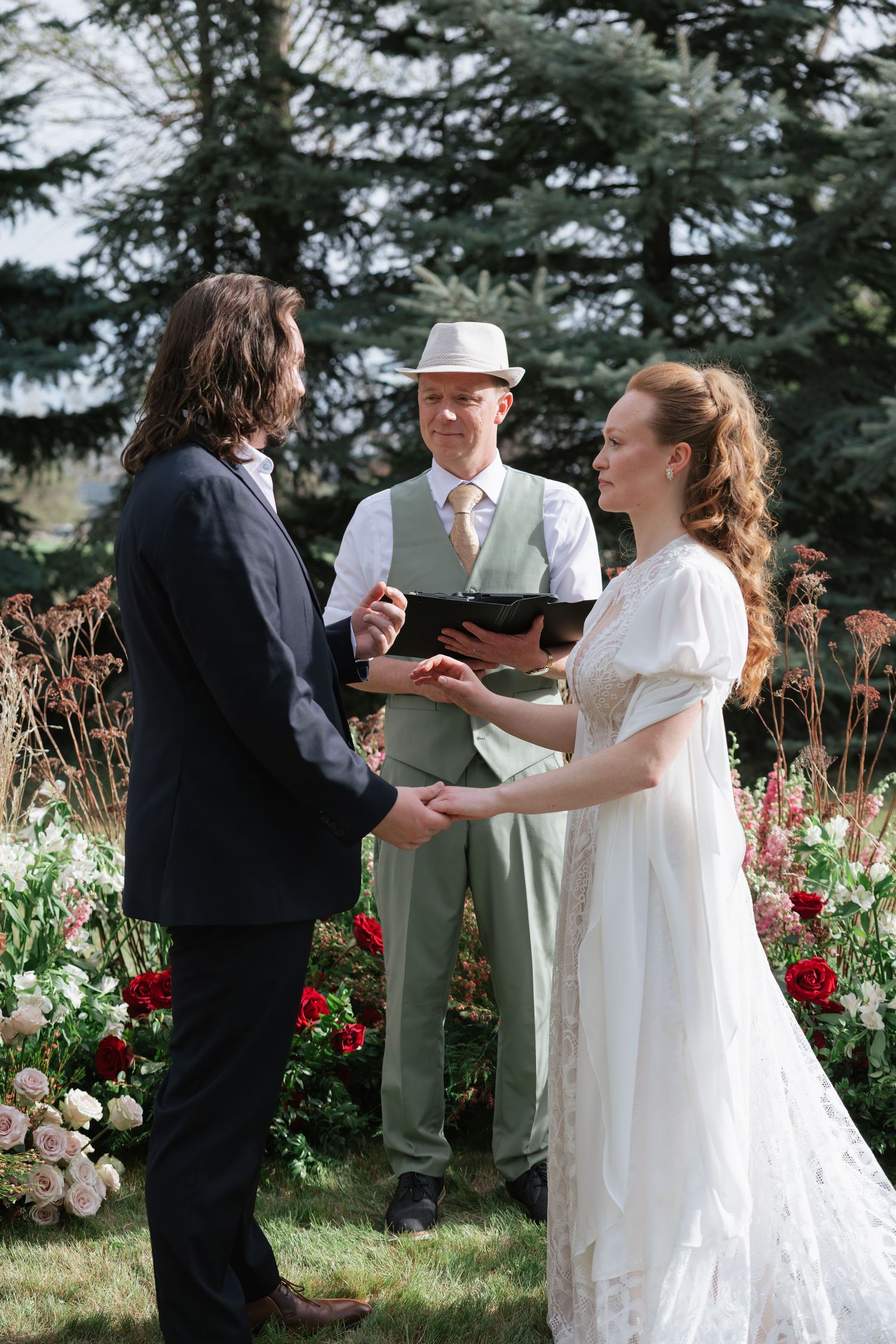 Bride and groom hold hands during outdoor wedding ceremony with officiant and floral backdrop.