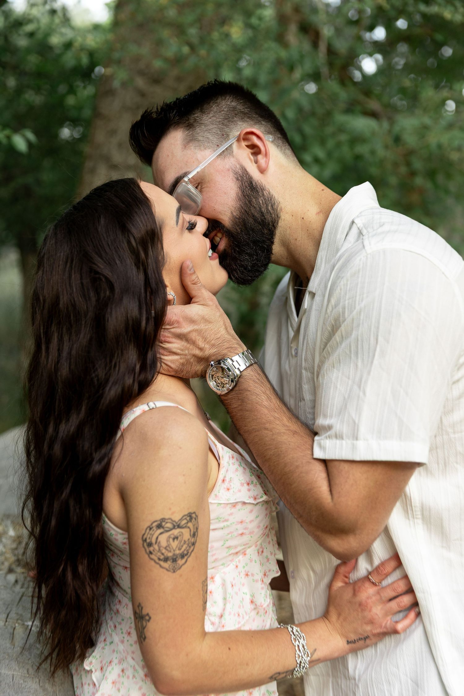 A bearded man in glasses tenderly kisses a brunette woman with a heart tattoo outdoors near green trees.