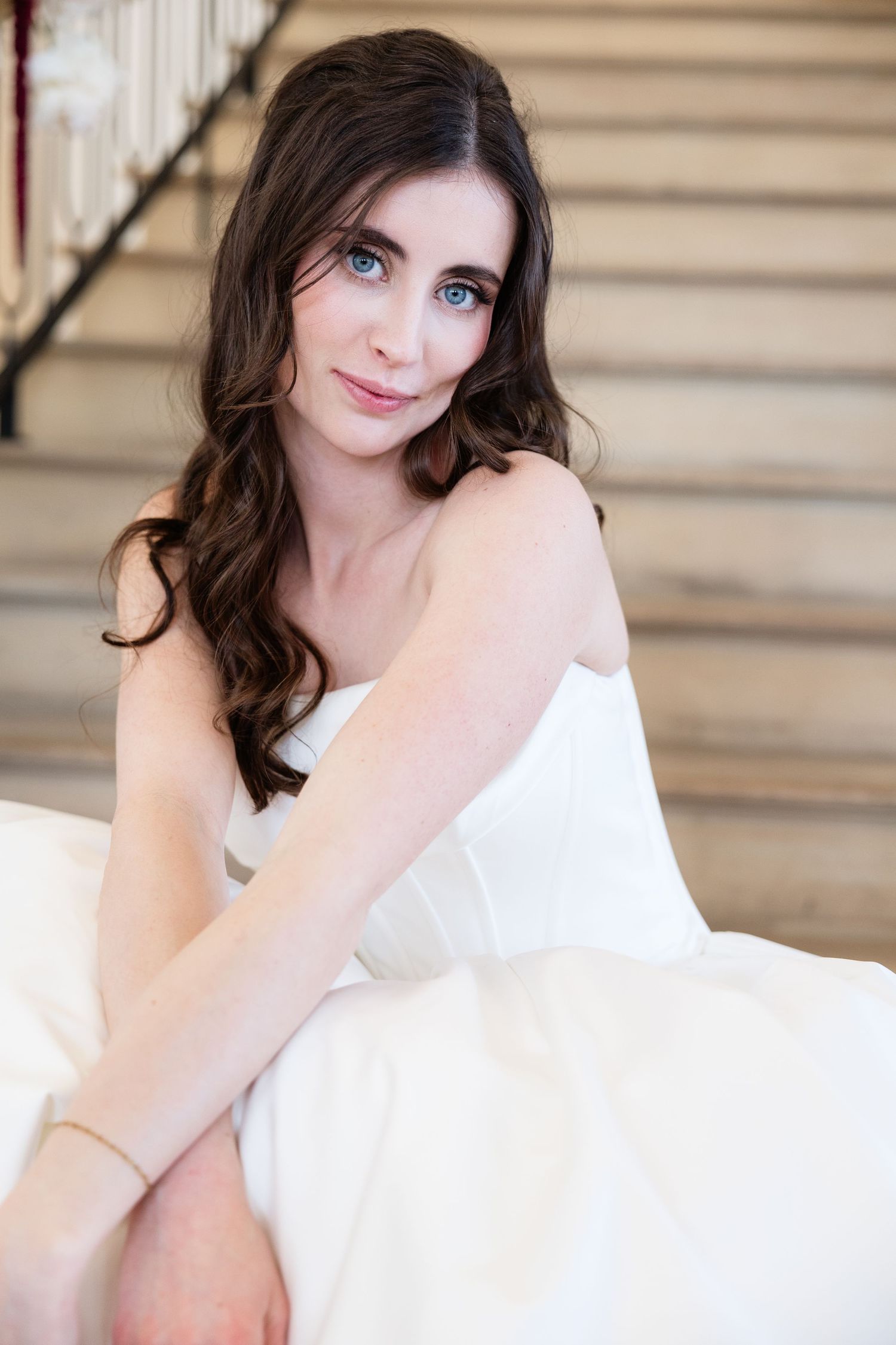 Elegant bride in white strapless gown sits on staircase steps, with blue eyes and long wavy brown hair.