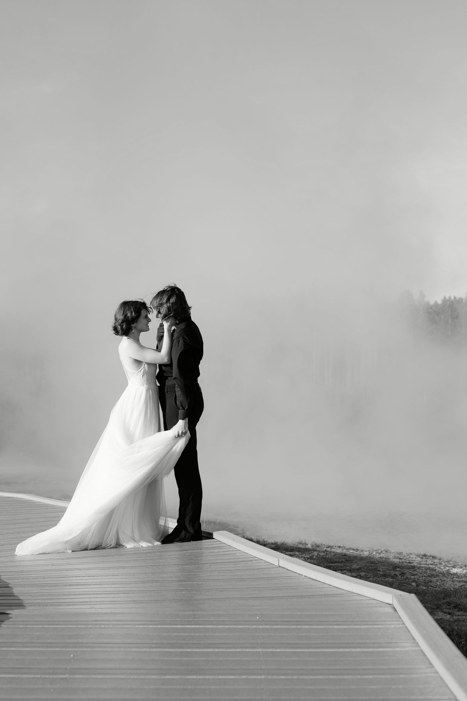 Black-and-white photo of a bride and groom embracing on the boardwalk at Grand Prismatic Spring in Yellowstone National Park,
