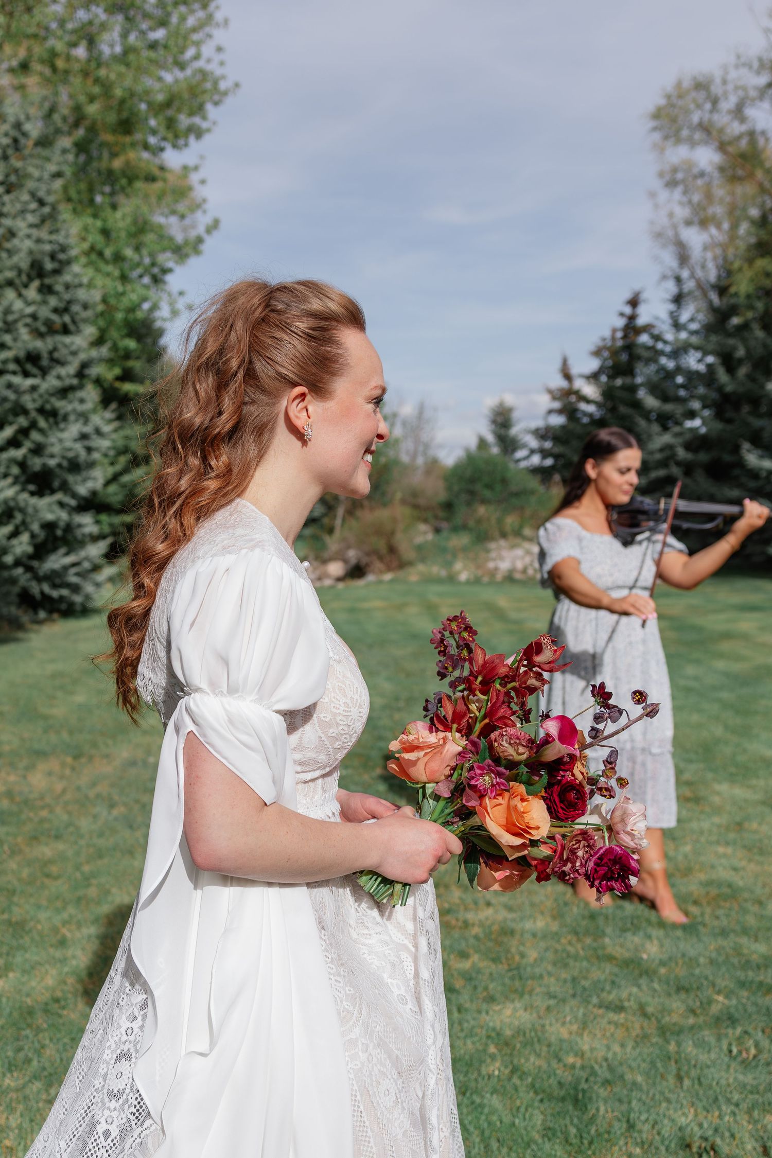 Smiling red-haired bride in white lace dress holding colorful bouquet while violinist plays outdoors.