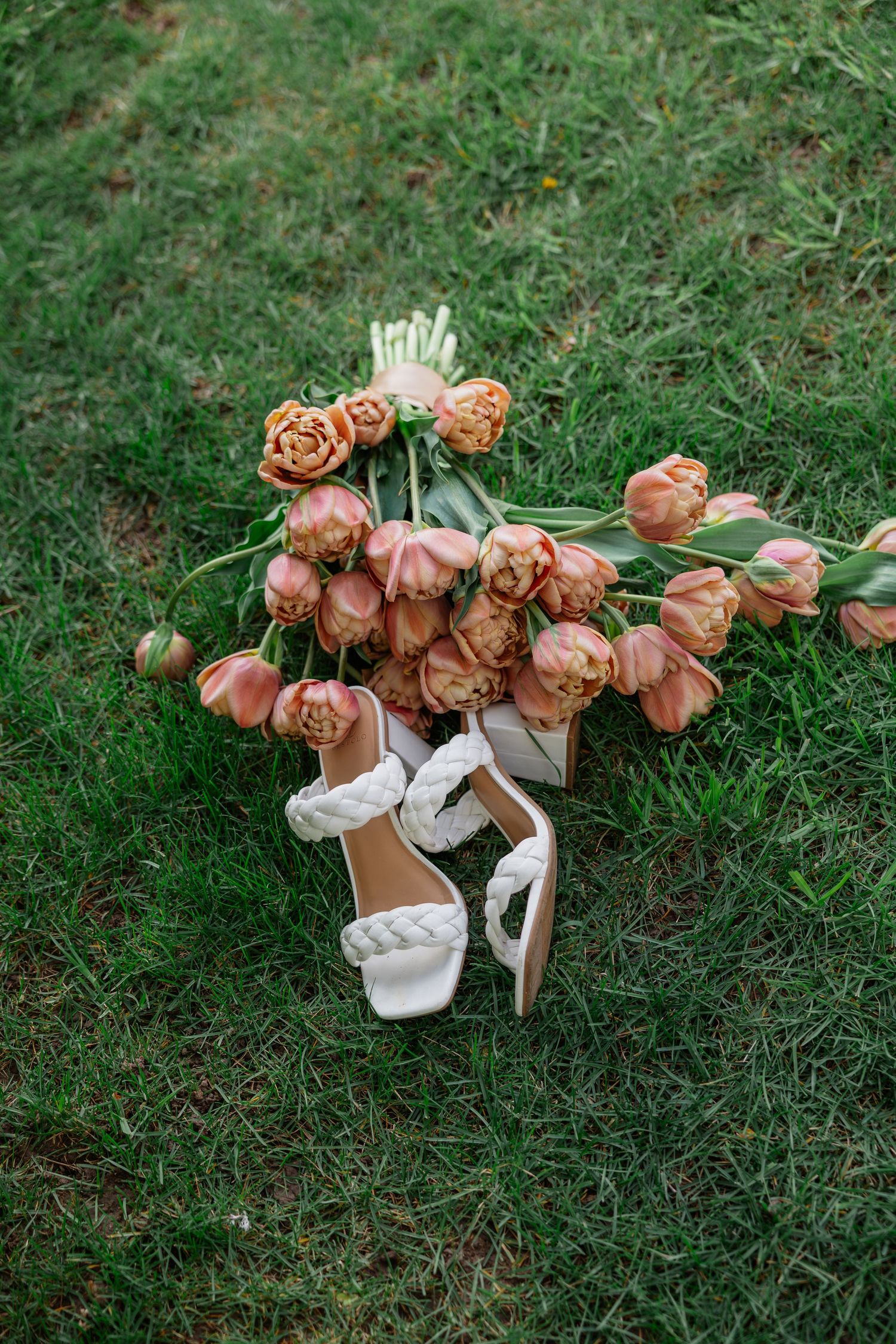 White braided block heel wedding sandals resting on green grass beside a bouquet of peach tulips.