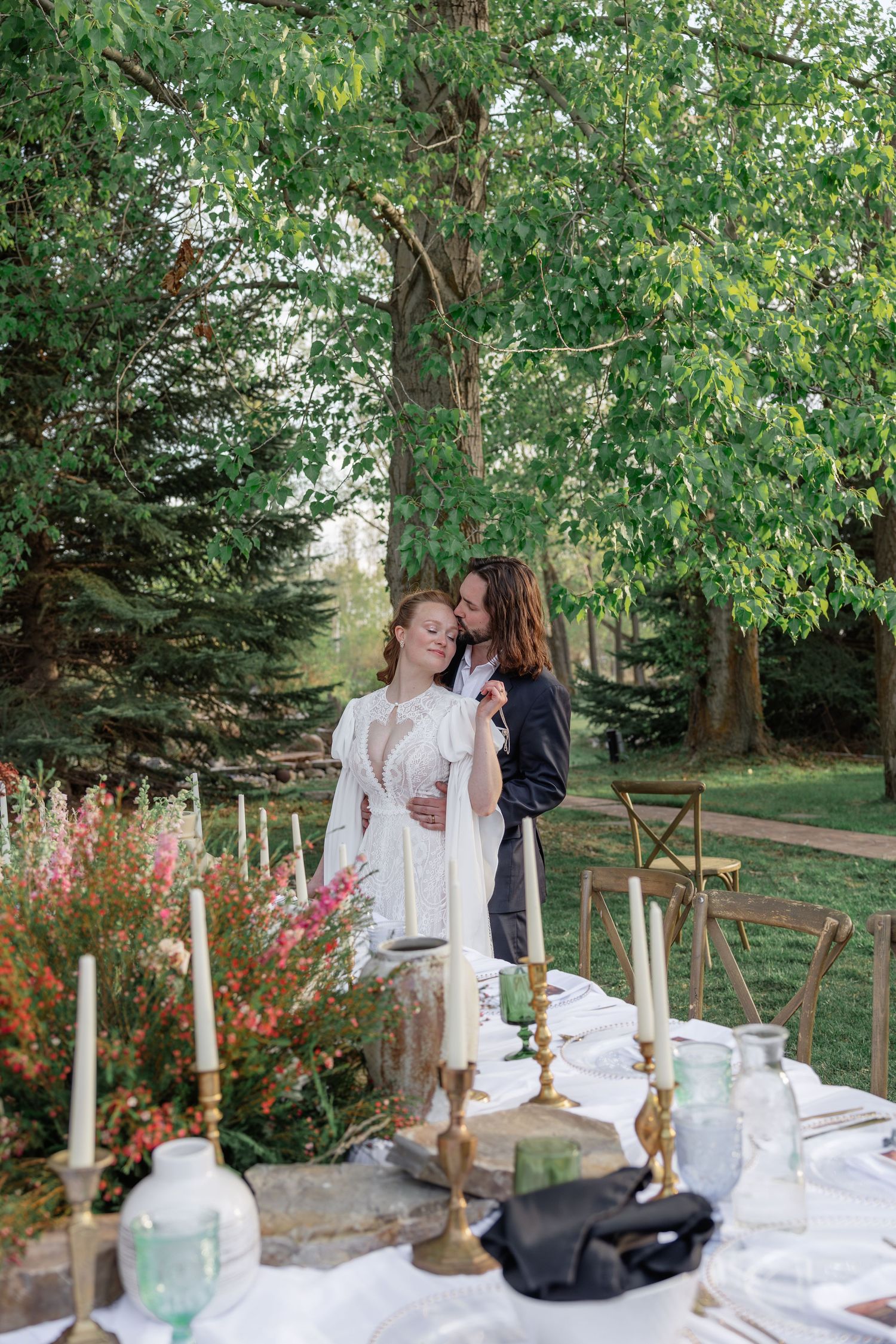 A couple embraces tenderly beside an elegantly set outdoor wedding reception table surrounded by lush greenery.