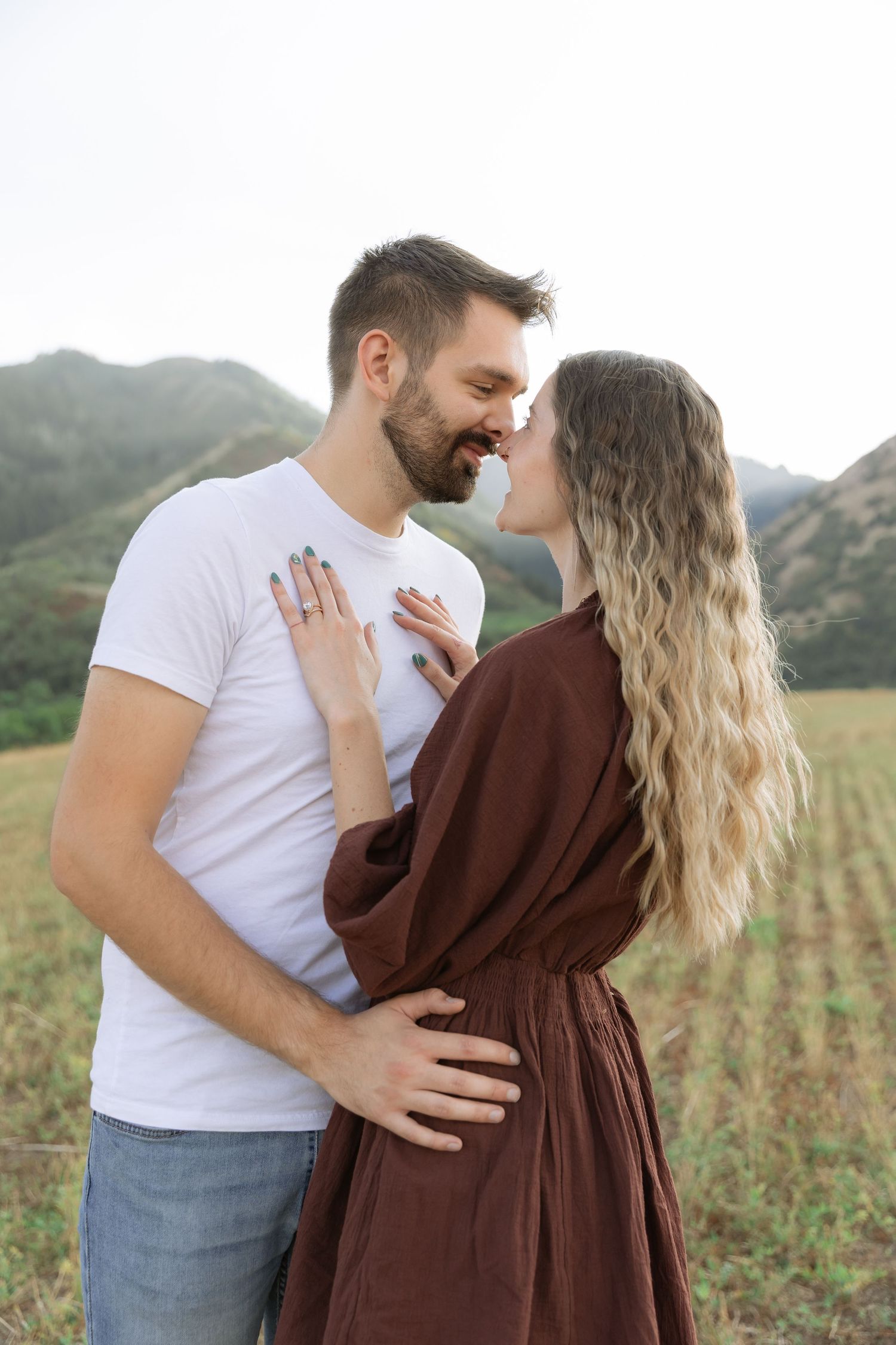 A couple sharing a romantic moment in a field with mountains, woman in brown dress, man in white t-shirt.