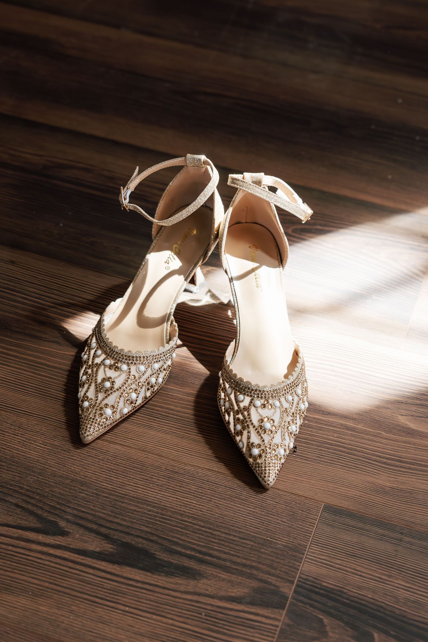 Elegant beaded bridal heels with ankle straps resting on dark hardwood floor in soft natural light.