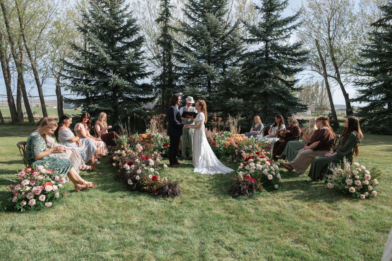 Small intimate outdoor wedding ceremony with guests seated in a circle around the couple amid tall pine trees and floral arrangements.