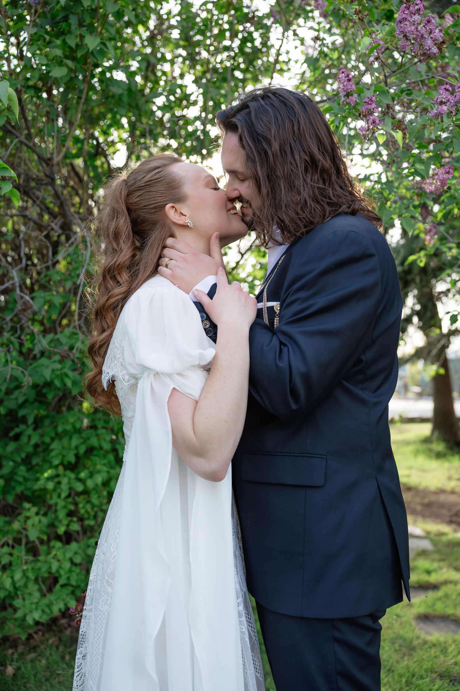 Bride and groom sharing a romantic kiss surrounded by green trees and blooming lilacs.