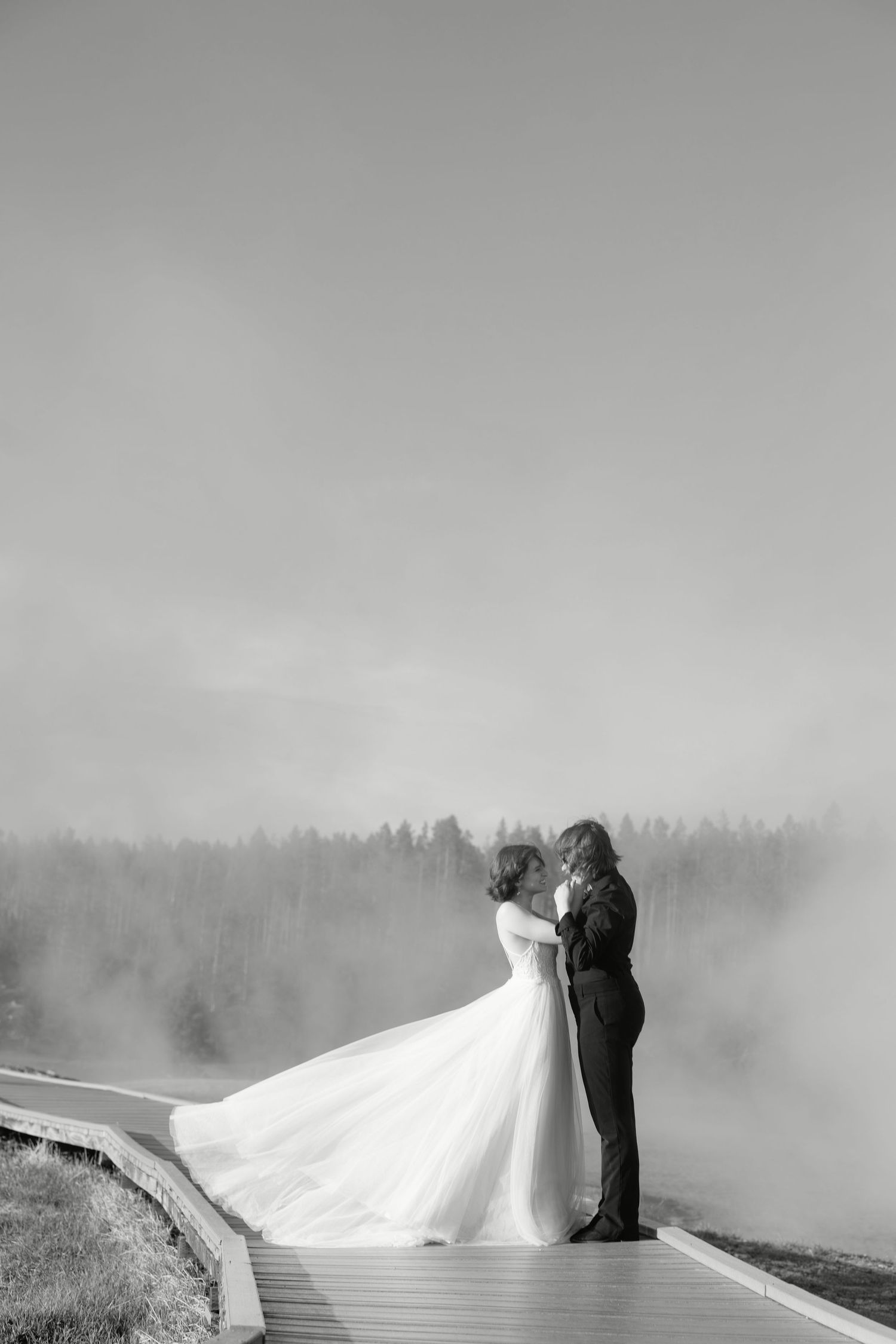 Black and white photo of bride and groom embracing on a misty boardwalk with foggy forest backdrop.